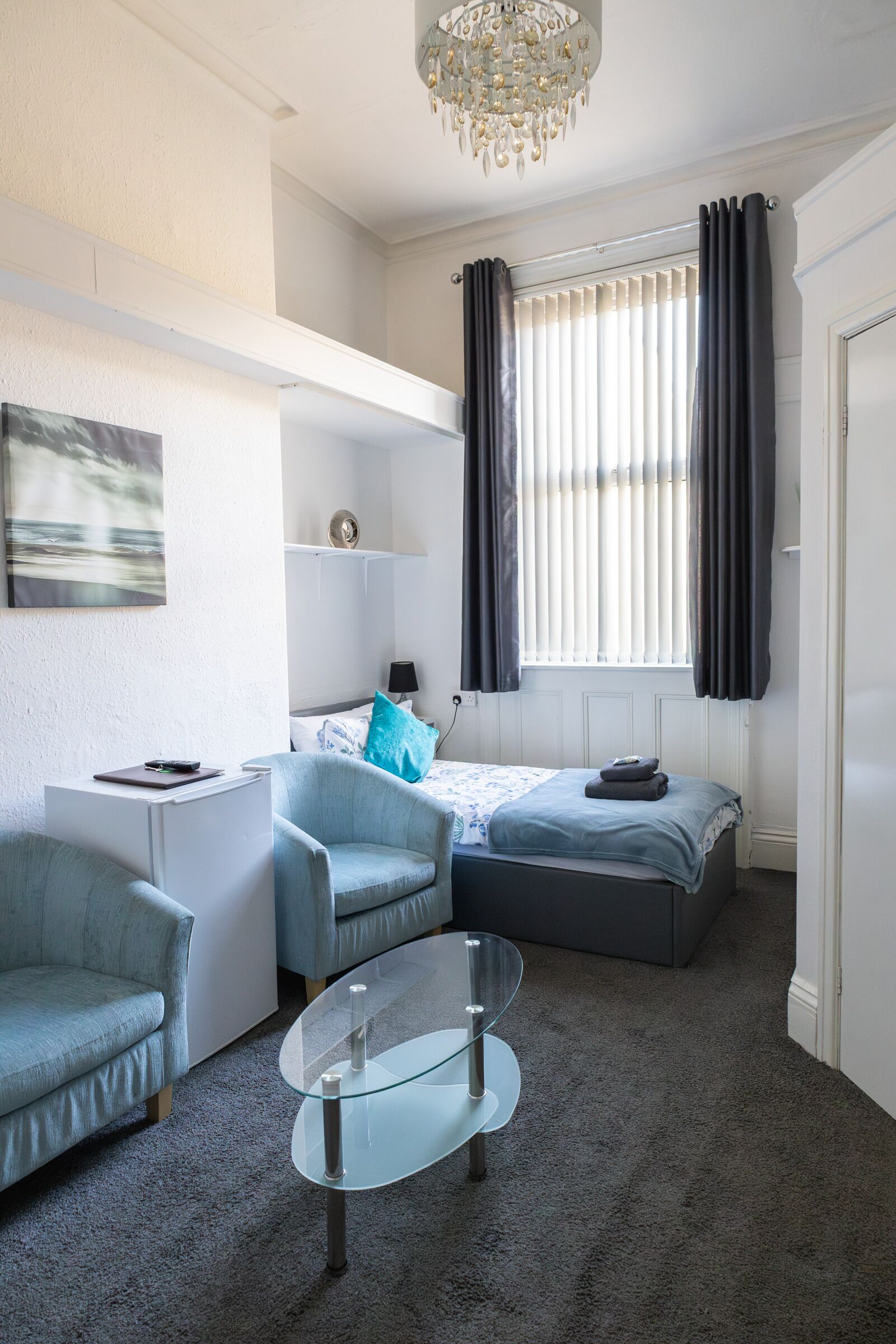 A compact room with a bed, a desk, a chair, a glass coffee table, and a chandelier.