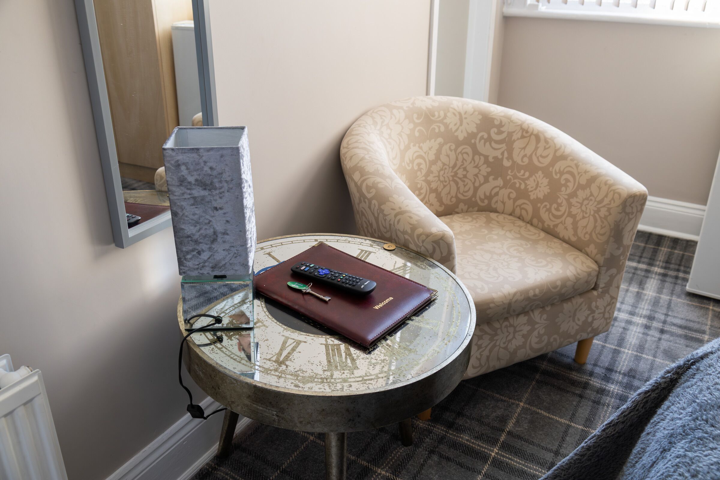A round glass table with a remote control, a notebook, and glasses. A patterned armchair is beside the table.