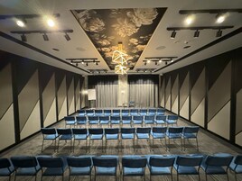 Un salón de conferencias con filas de sillas, un escenario, un diseño floral en el techo y una lámpara moderna.