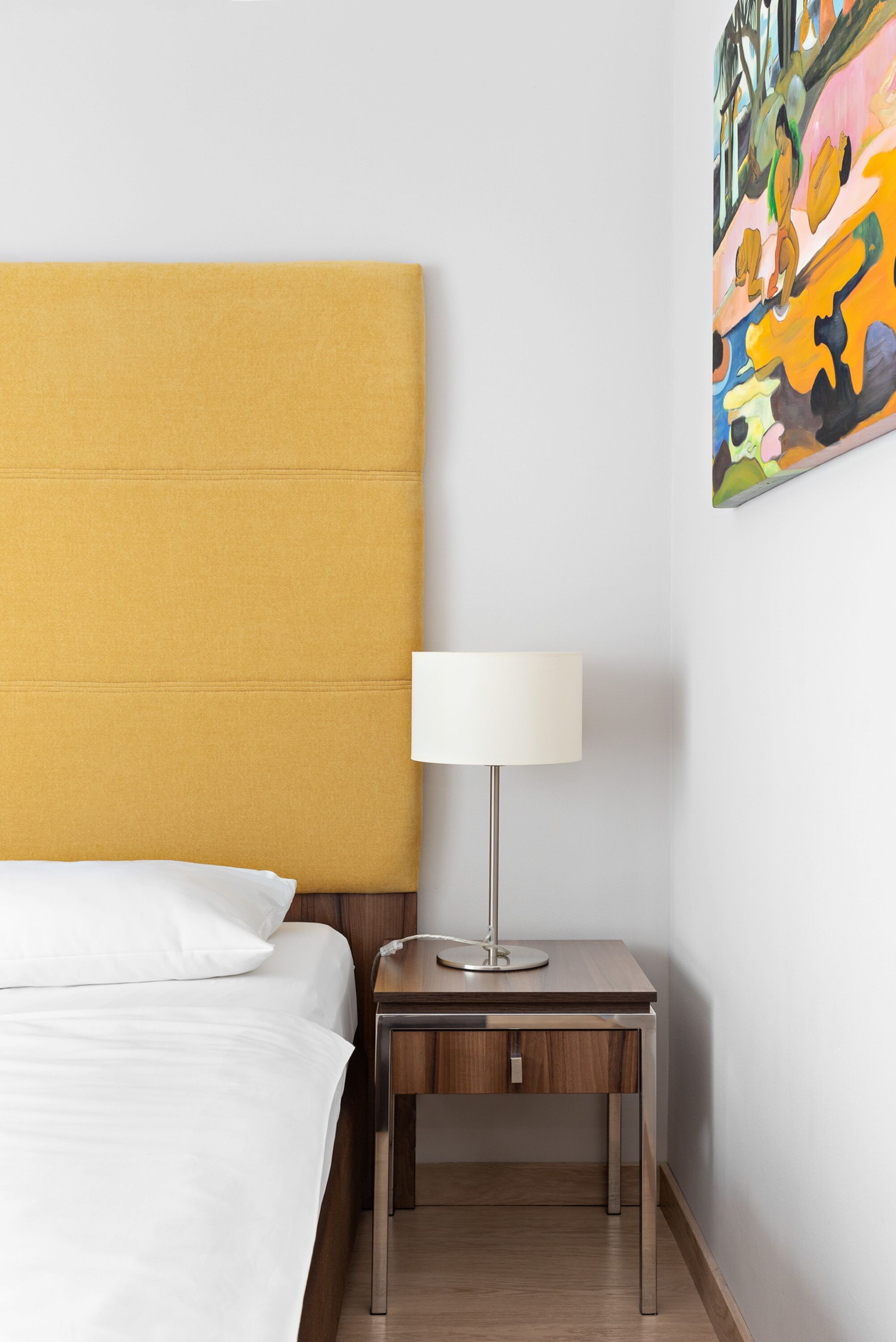 A hotel room with a bed, a nightstand with a lamp, and a colorful abstract painting on the wall.