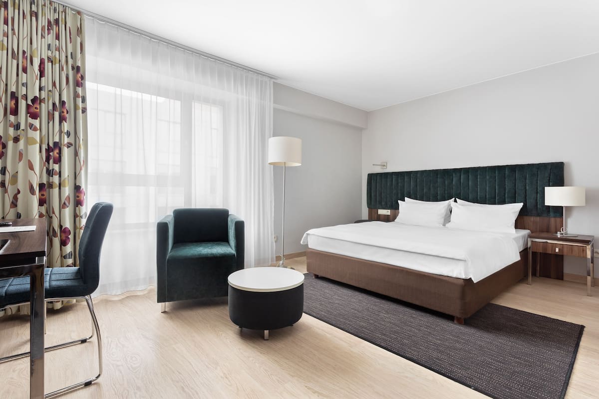 A hotel room with a bed, a desk, a chair, a lamp, and a large window with sheer curtains.
