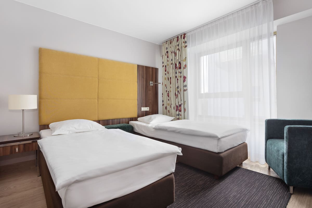 A hotel room with two beds, a desk, a chair, and a large window with sheer curtains.
