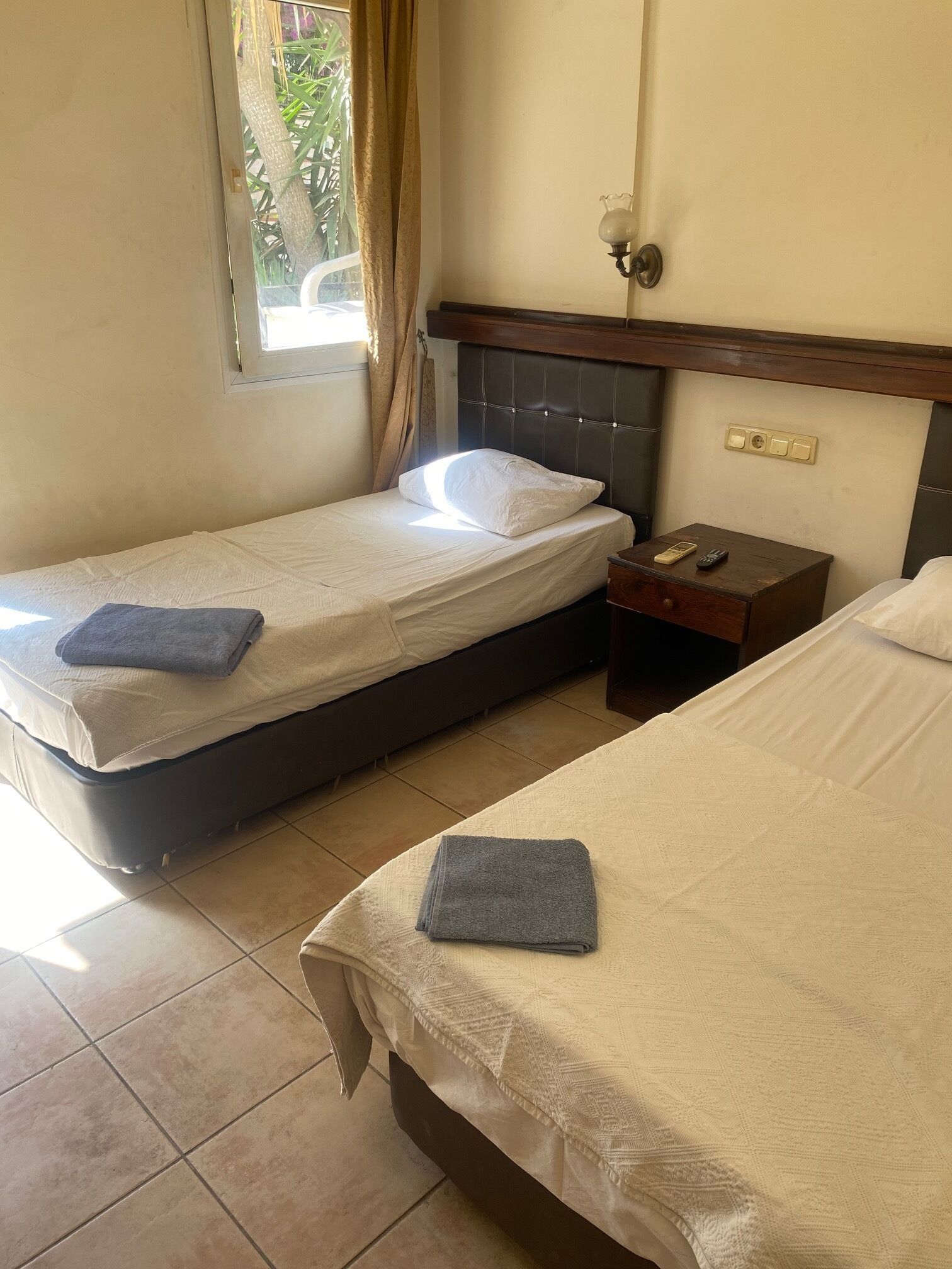 Une chambre d’hôtel avec deux lits, une table de chevet, une fenêtre avec des rideaux et un luminaire fixé au mur.