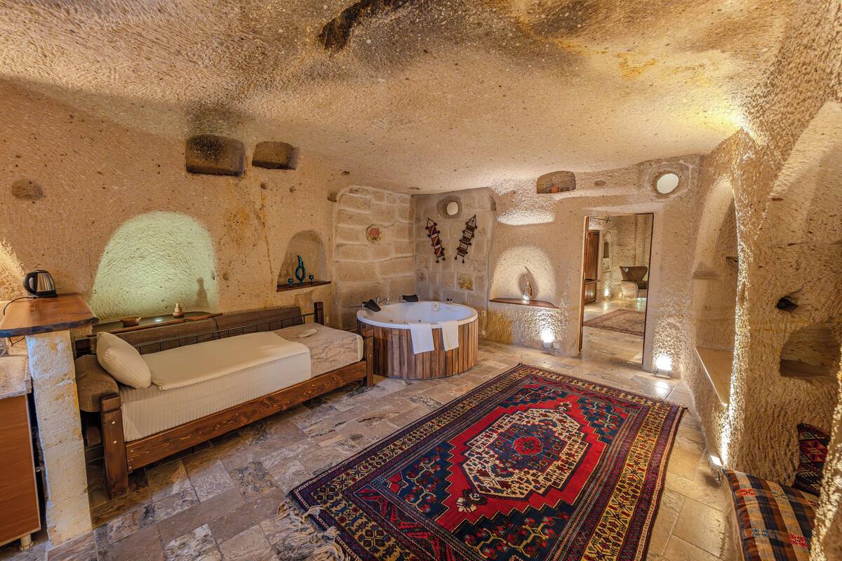 A stone-walled room with a wooden bed, a round bathtub, and a red patterned rug.