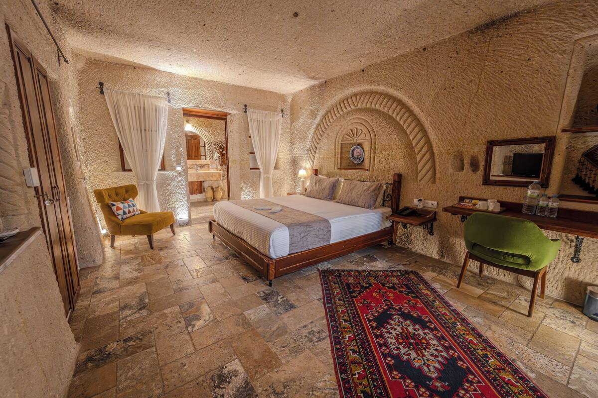 A bedroom with a bed, a chair, a nightstand, a green chair, a rug, and a stone wall.