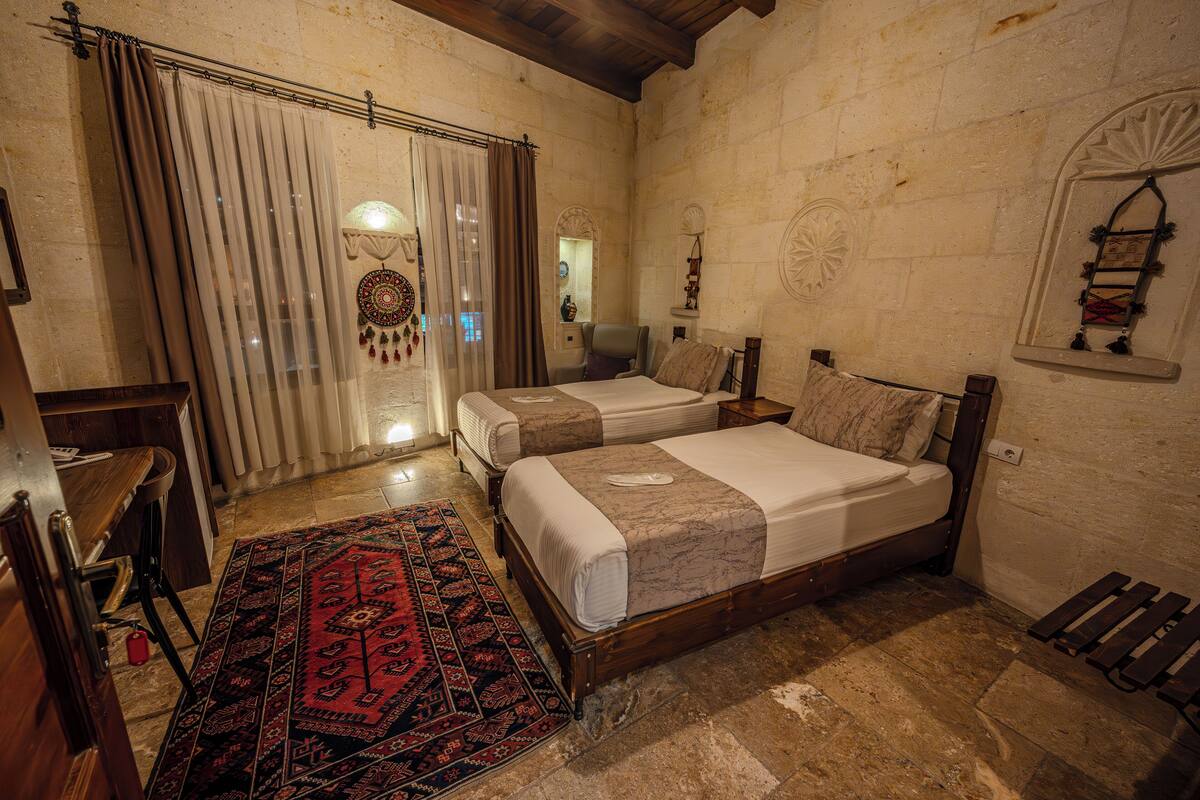 A room with two beds, a wooden bench, a red patterned rug, and stone walls.