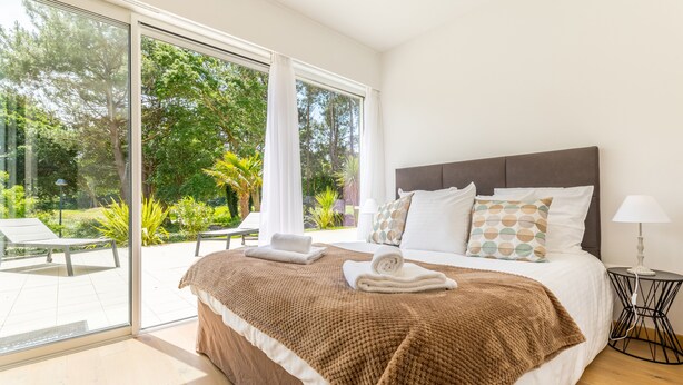 A bedroom with a large bed, a bedside table with a lamp, and a view of a garden through a sliding glass door. - Madame Vacances Les Palmiers du Golf (Moliets-et-Maa)