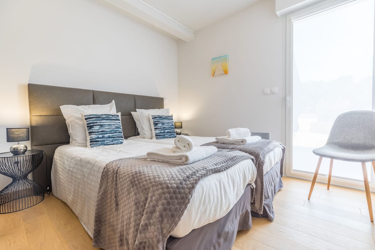 A modern hotel room with a large bed, two bedside tables, a chair, and a window with a view.