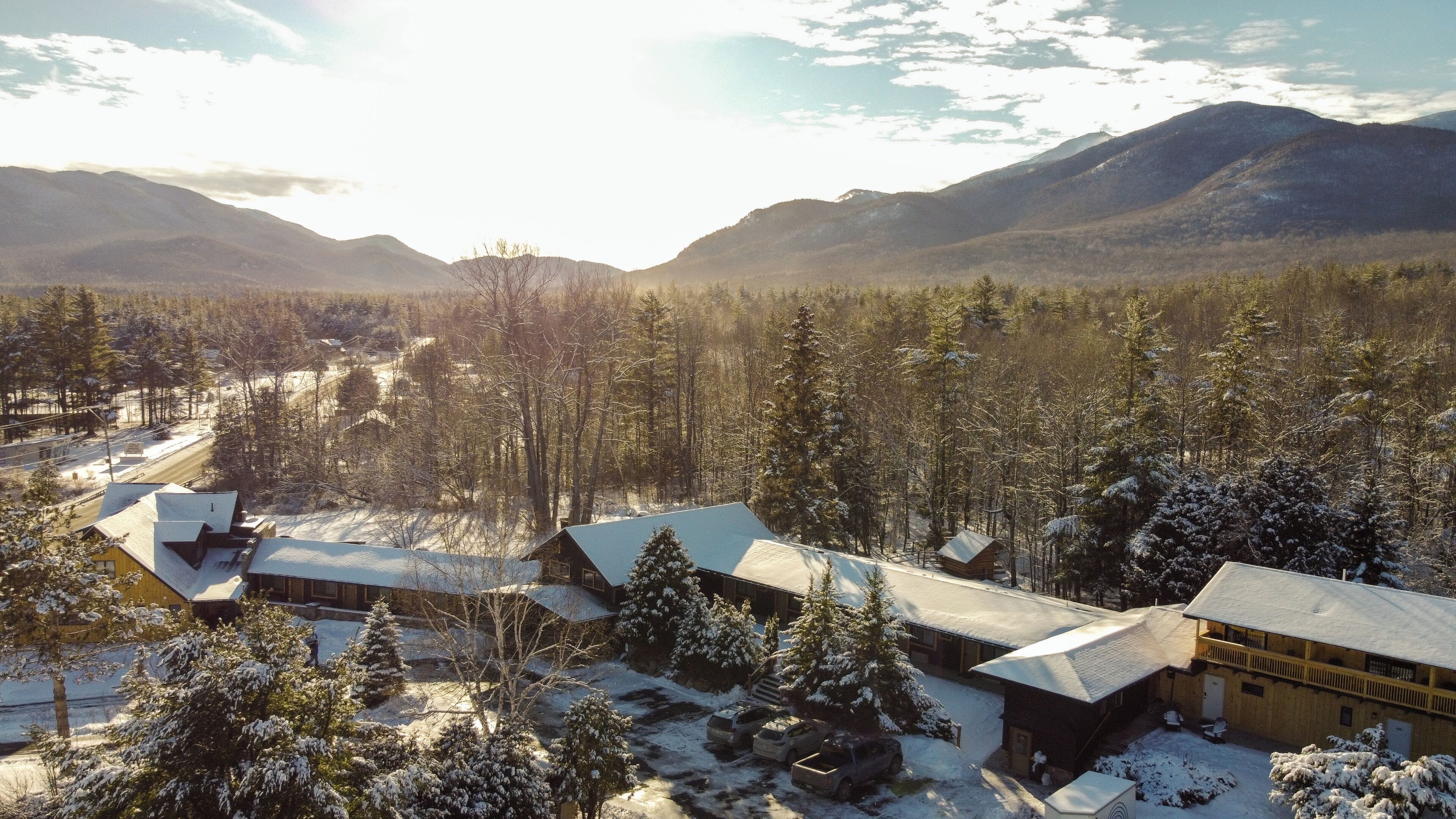 A cabin with a snow-covered roof, surrounded by a forest and mountains.