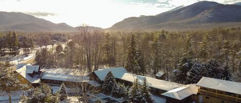 A cabin with a snow-covered roof, surrounded by a forest and mountains.