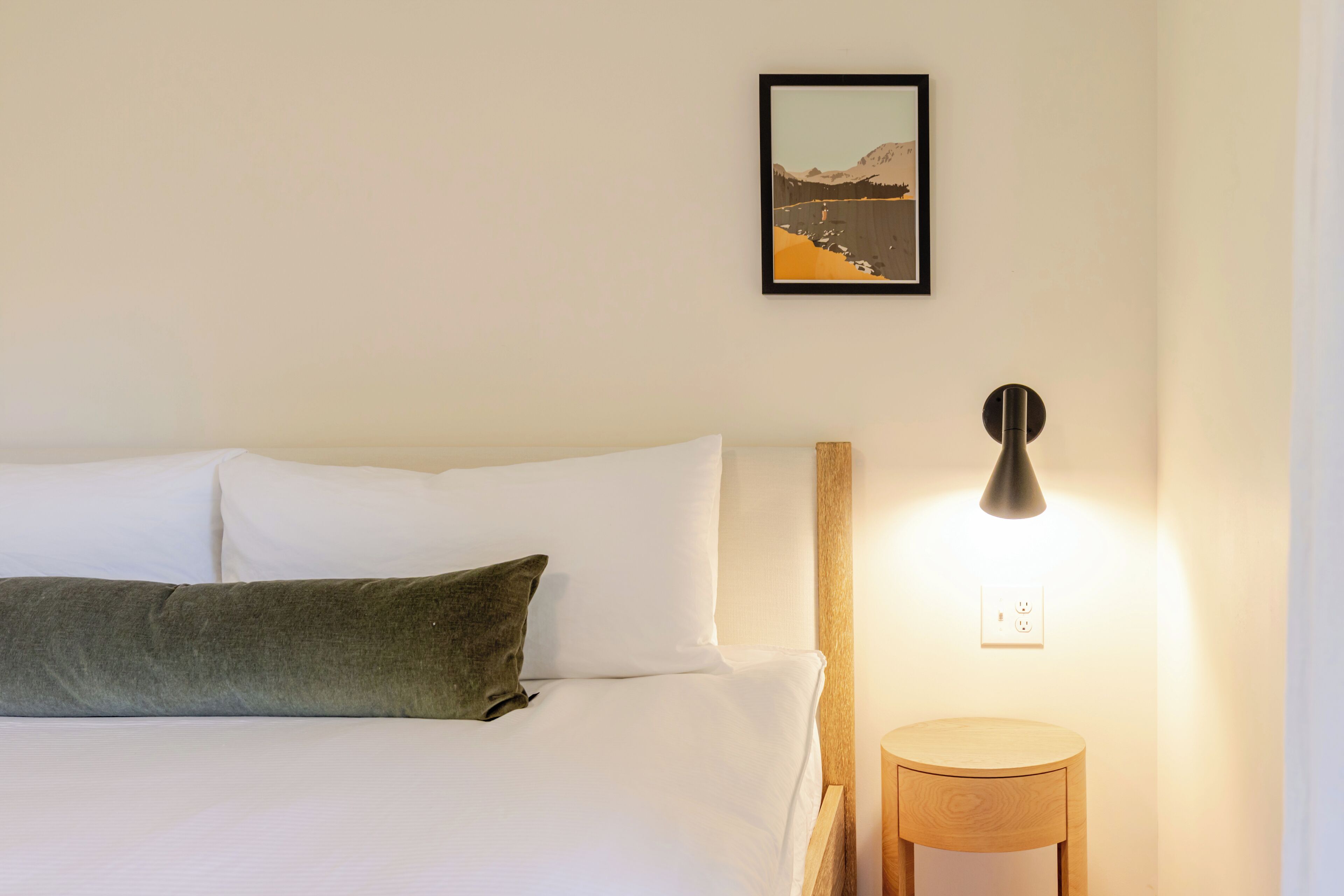 Ein ordentlich bezogenes Bett mit weißen Leinen und einem grünen Kopfkissen, ein gerahmtes Bild an der Wand, ein runder Nachttisch mit einer Lampe und eine Steckdose.