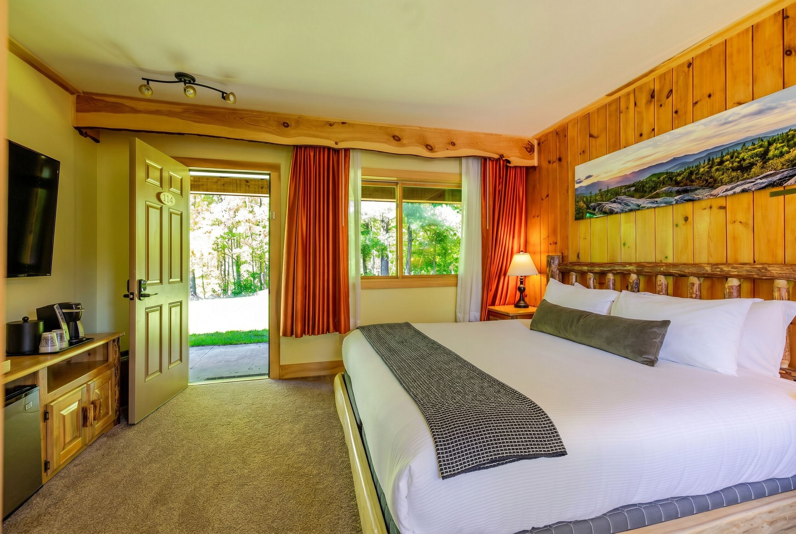 A hotel room with a bed, a desk with a coffee maker, a painting on the wall, and a view of greenery outside.