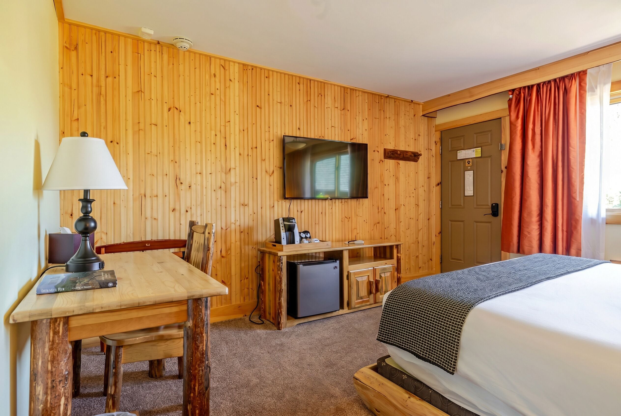 A room with a wooden wall, a television mounted above a wooden desk, a bed with a blanket, and a door with a sign.