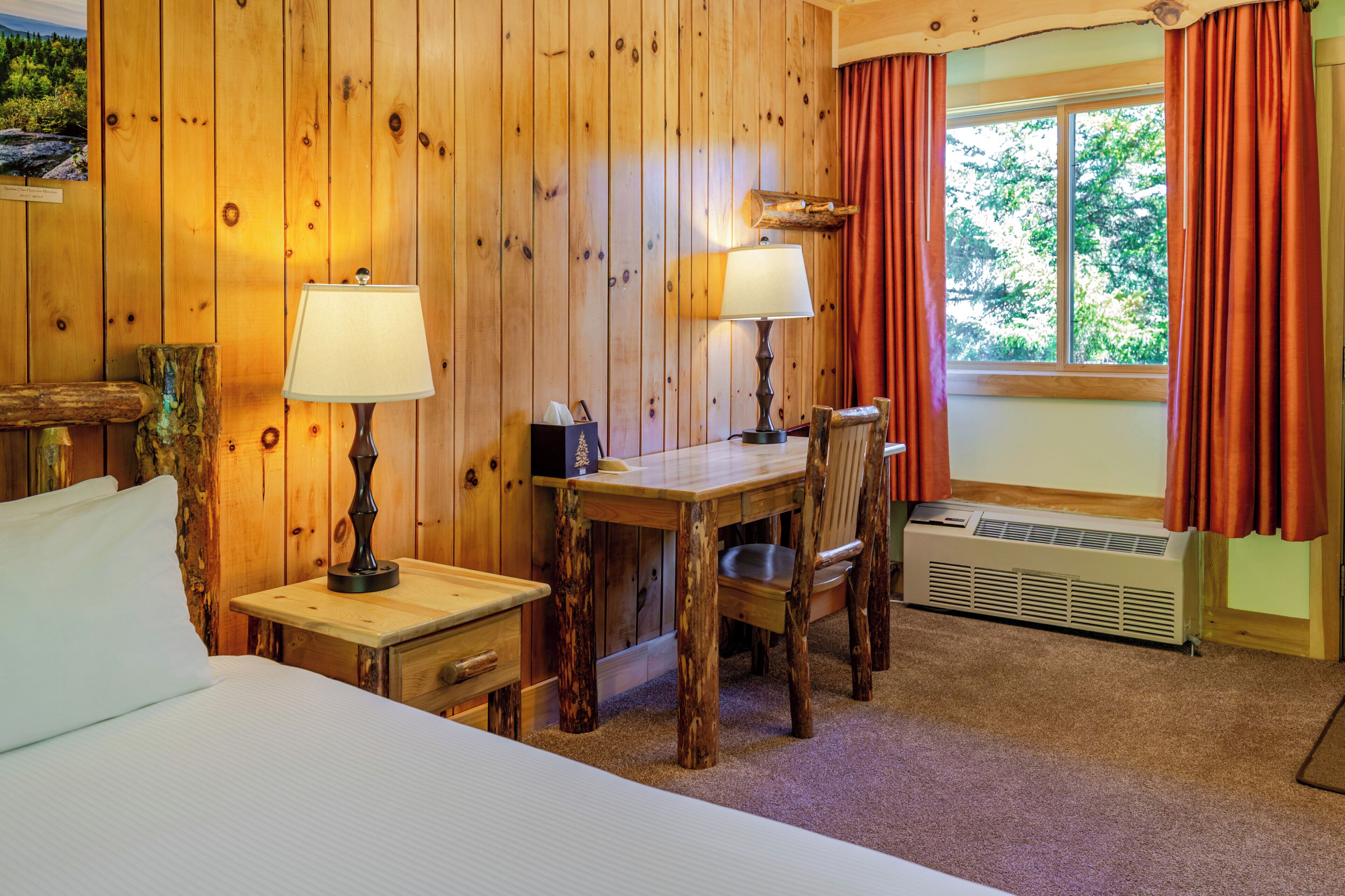 A wooden room with a bed, a table, two chairs, a window with curtains, and a wall-mounted heater.