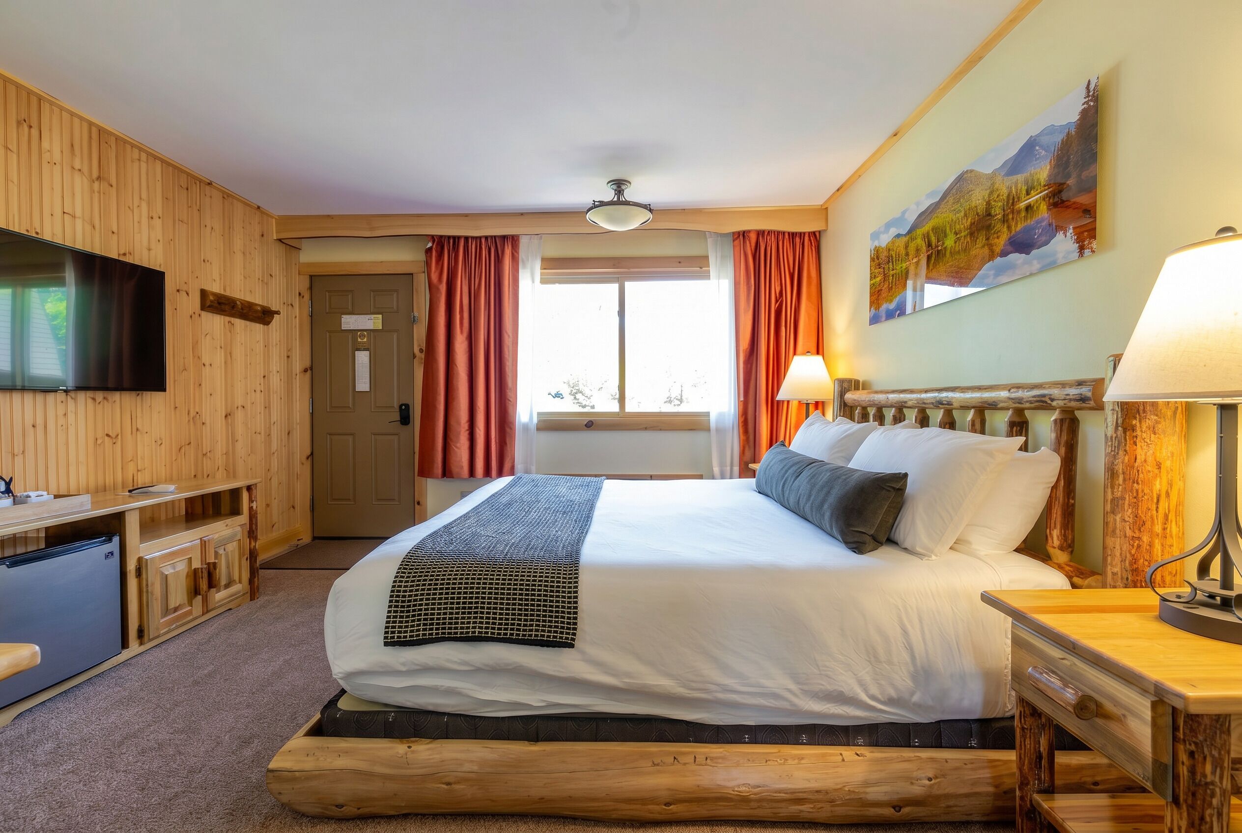 A wooden-themed hotel room with a bed, desk, TV, and a painting of a mountain landscape.