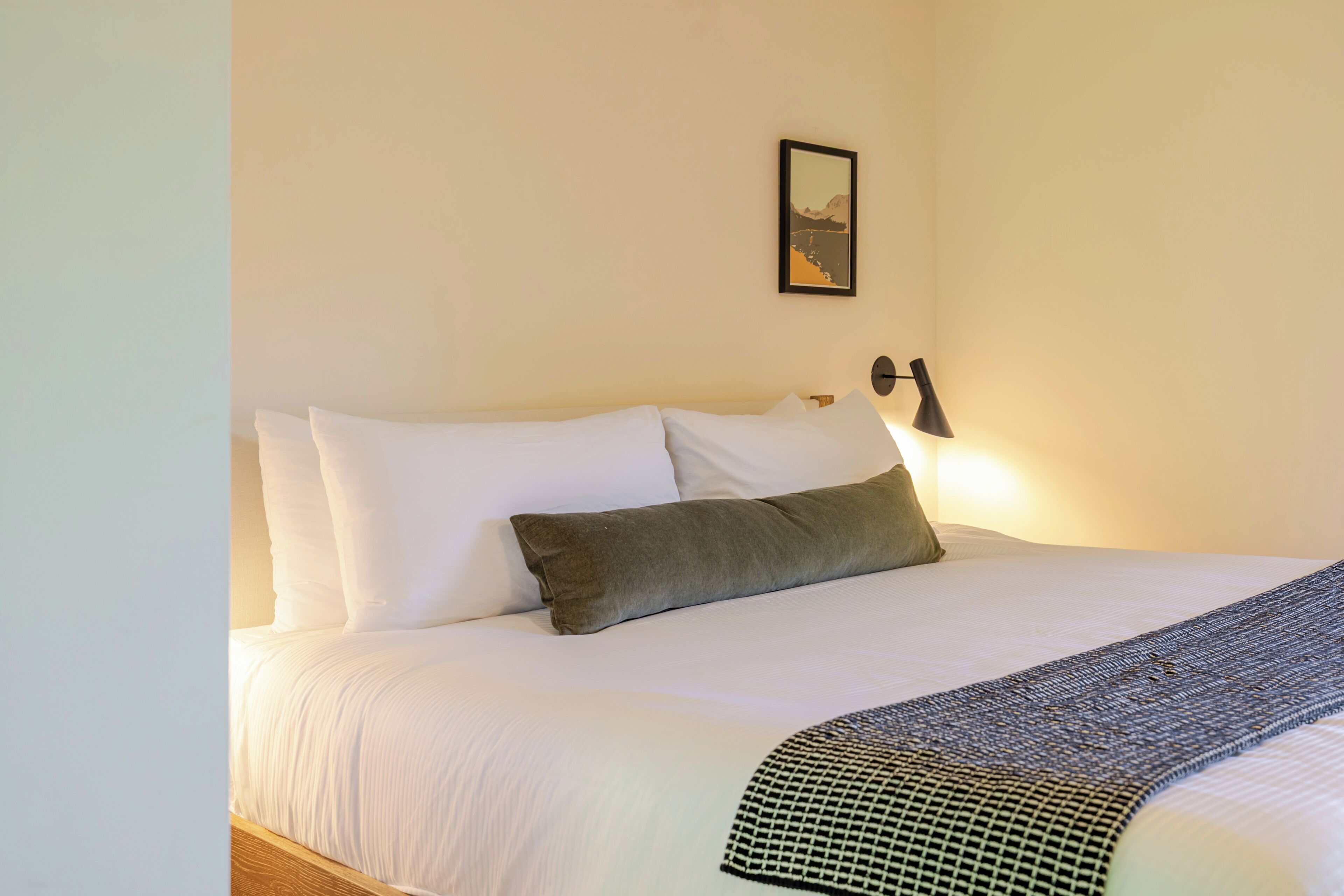 A neatly made bed with white and gray bedding, a framed picture on the wall, and a wall-mounted lamp.