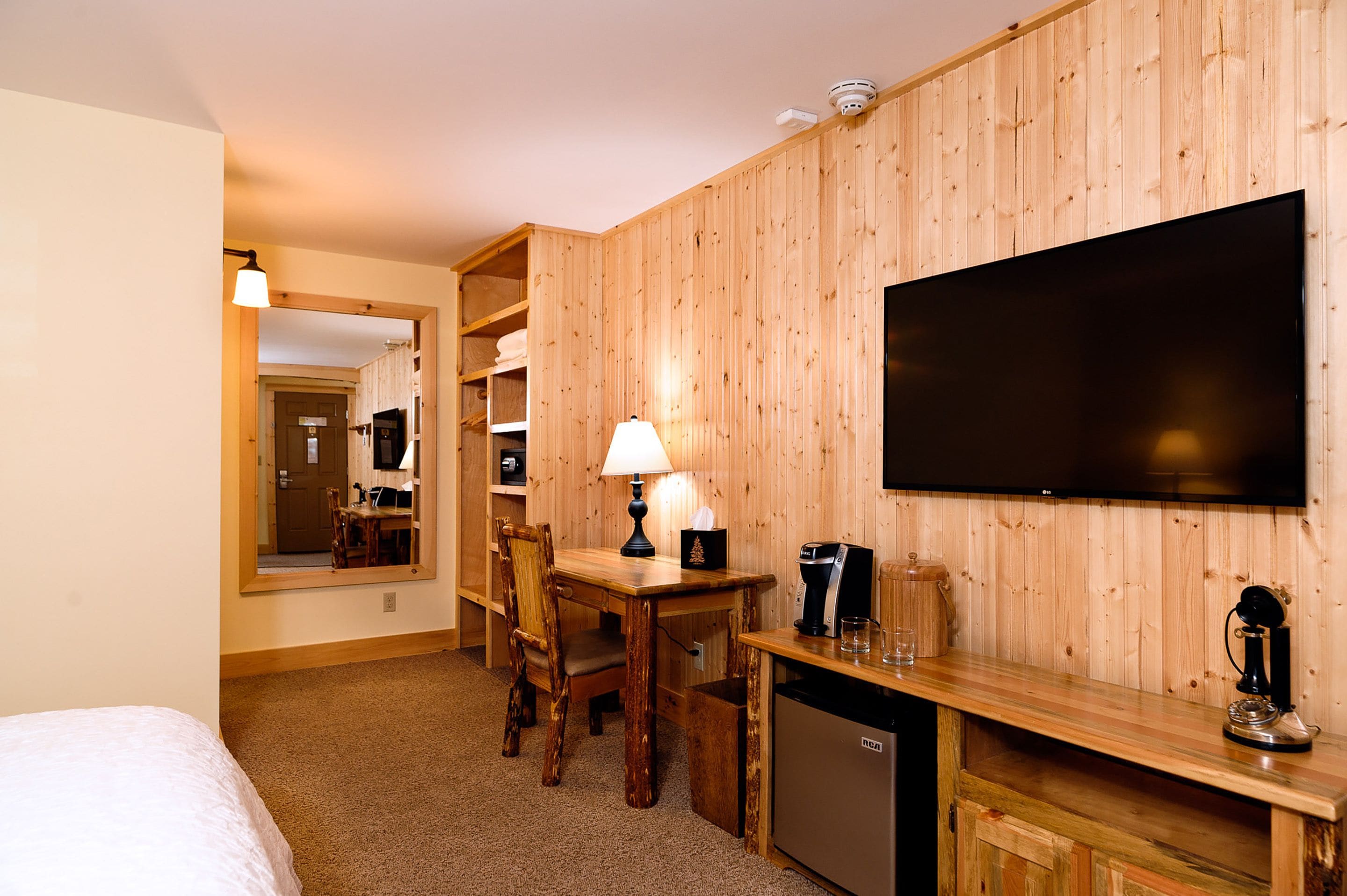 A hotel room with a wooden wall, a flat-screen TV, a wooden desk with a lamp, a coffee maker, and a mini-fridge.