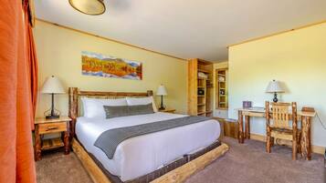 A room with a wooden bed, two bedside tables with lamps, a desk with a chair, a bookshelf, and a painting on the wall.