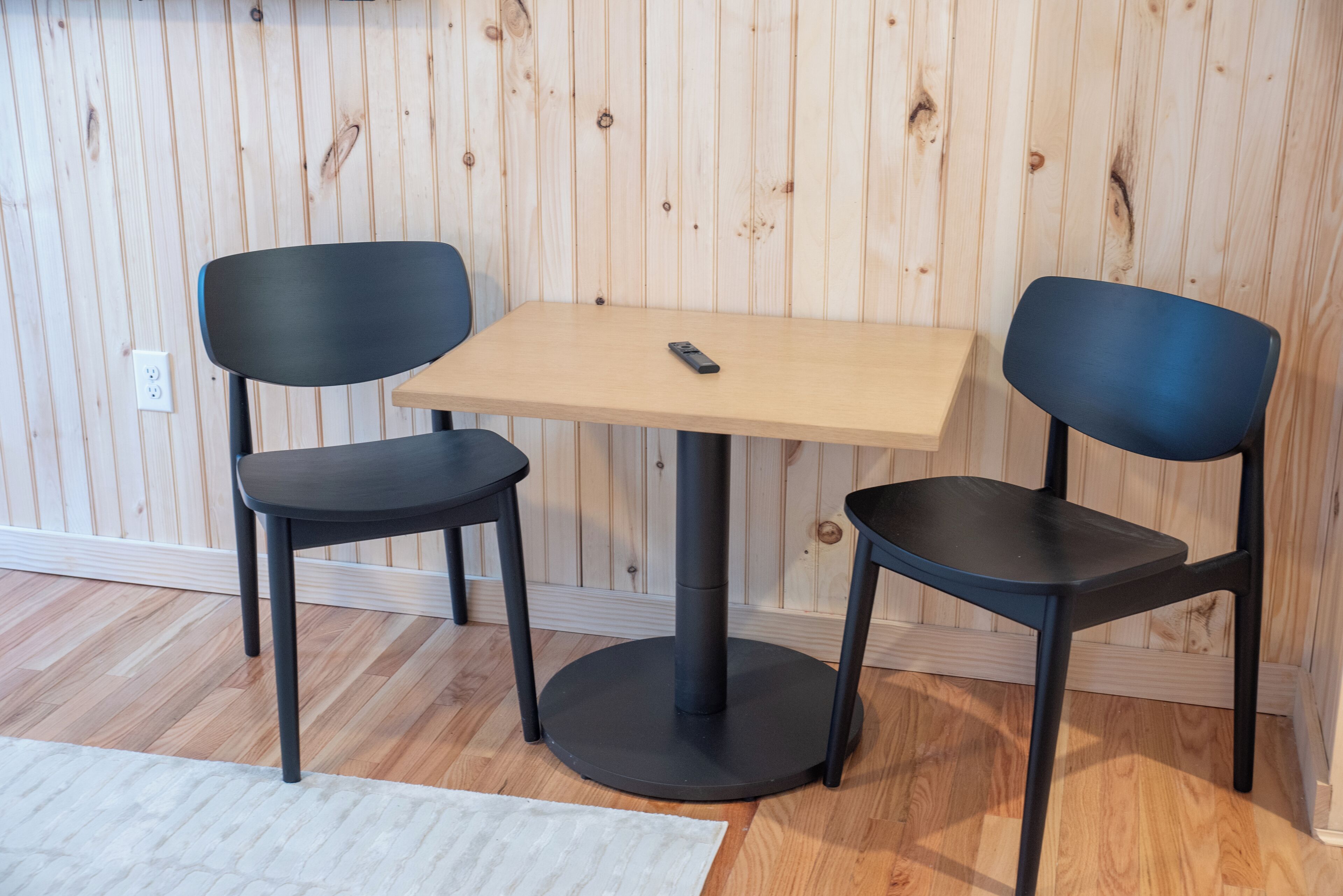 A room with a wooden paneled wall, a round table, two black chairs, and a white rug.