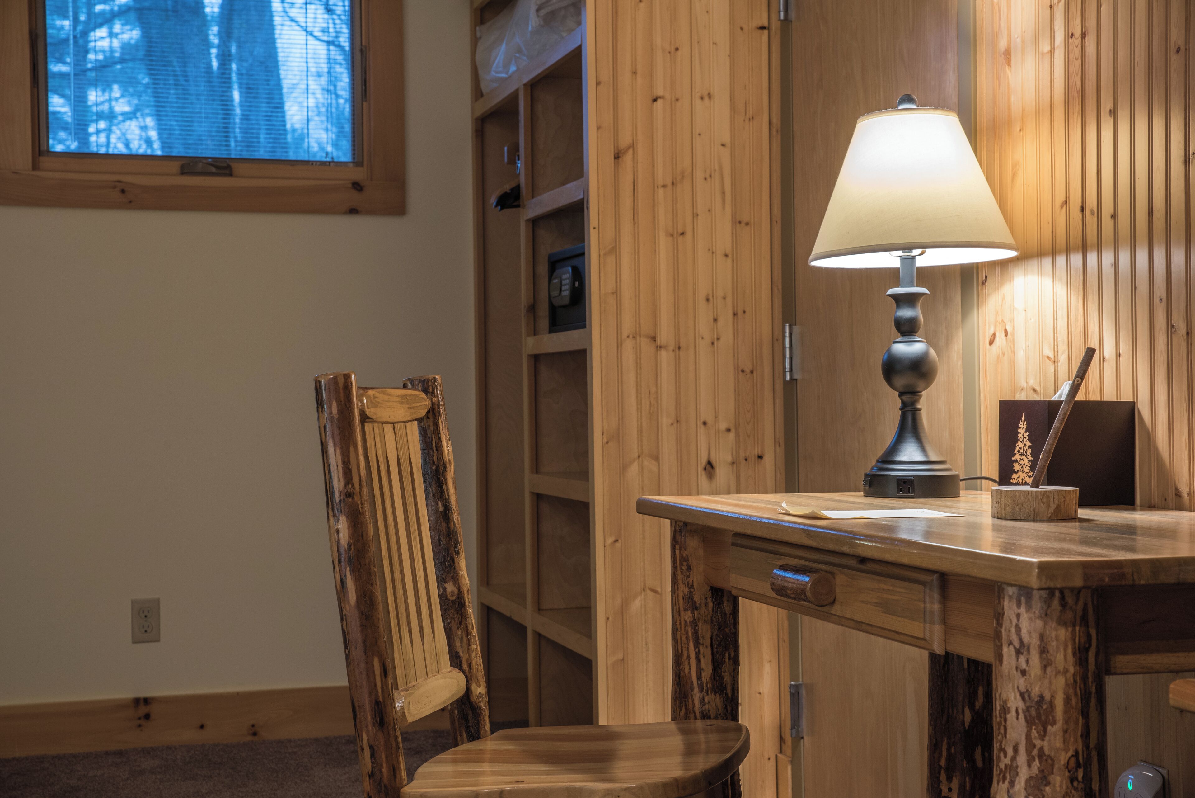 Un escritorio de madera con una lámpara, una silla y un ventanal con vista a árboles.