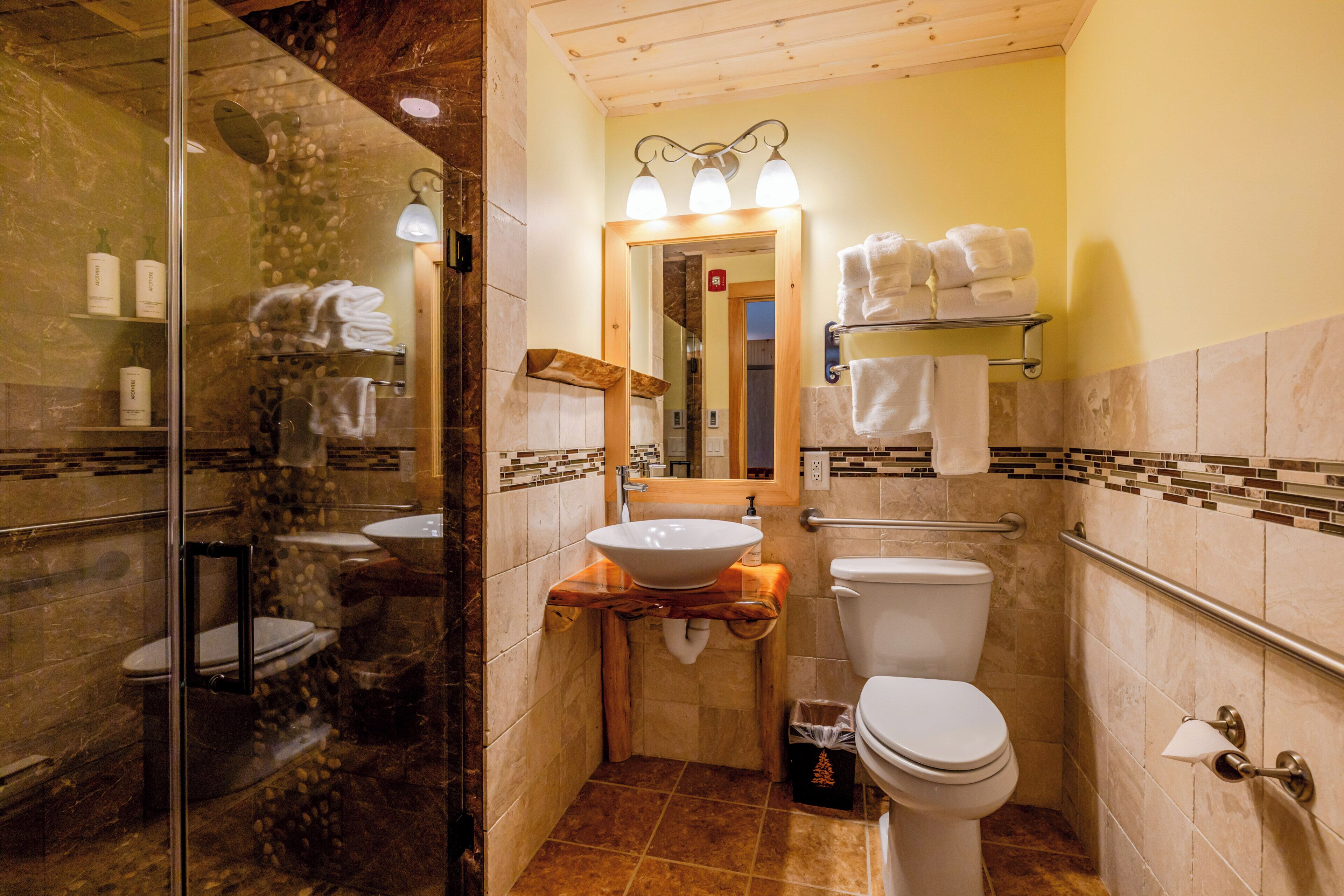 A bathroom with a glass shower enclosure, a sink with a vessel basin, a toilet, and a mirror with a towel rack above it.