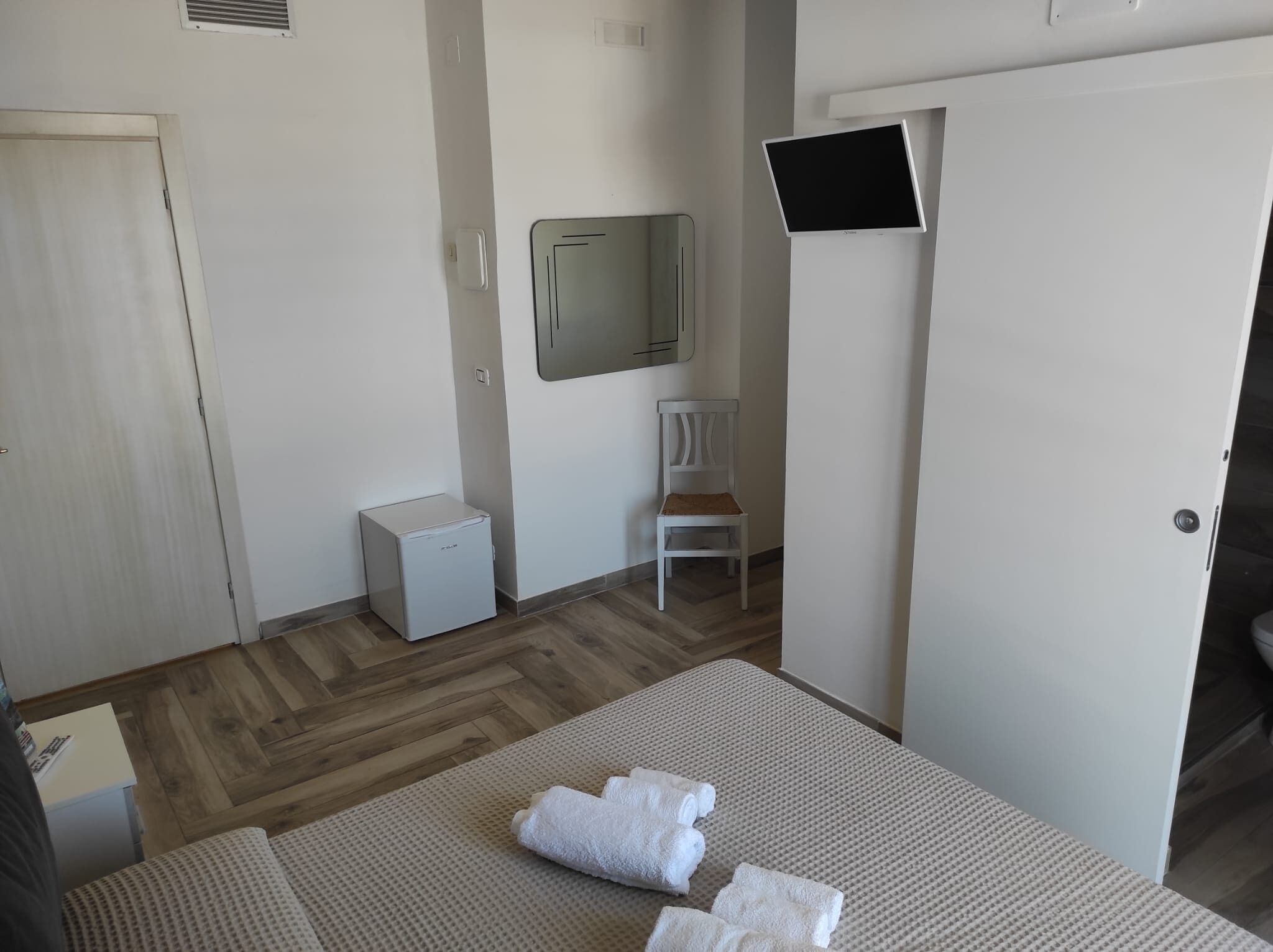 A hotel room with a bed, a small white chair, a television mounted on the wall, and a compact refrigerator.