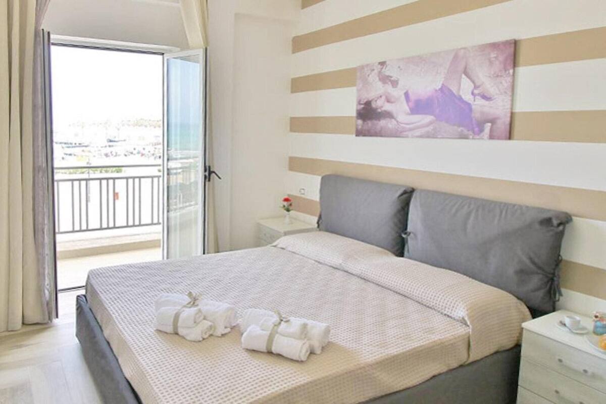 A hotel room with a bed, a nightstand, a painting, and a view of the city through the balcony.