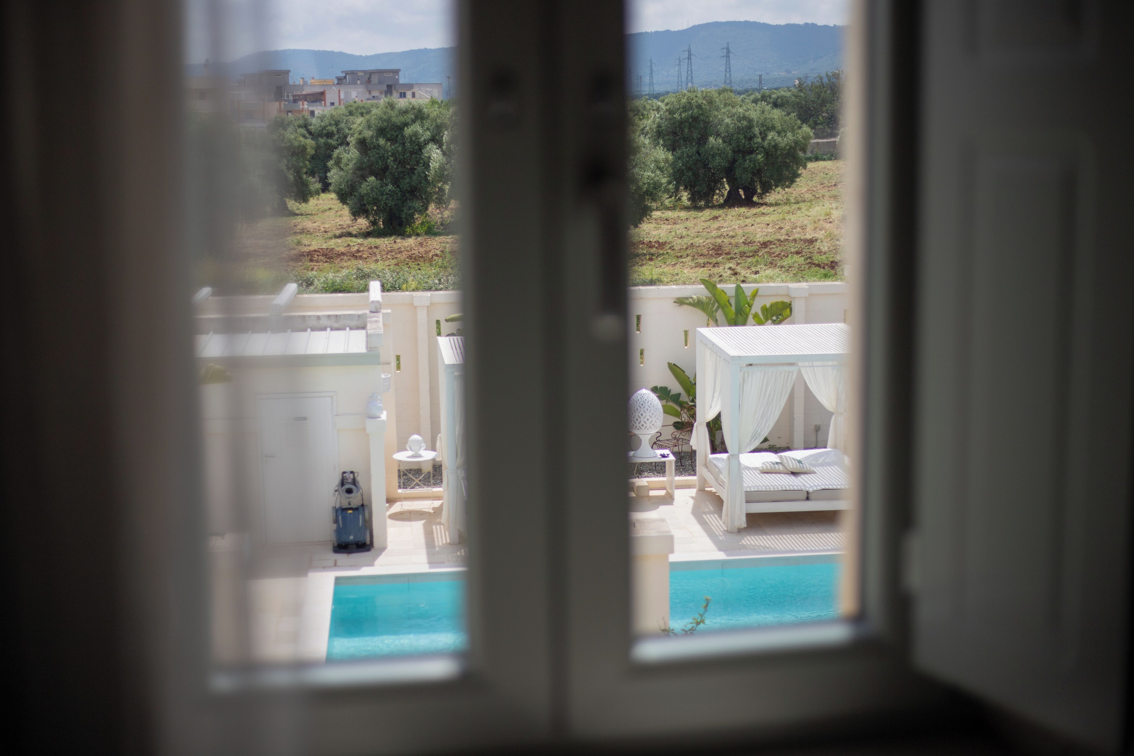 Une vue depuis la fenêtre d’une chambre donnant sur une piscine, un lit et une plante.