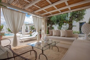 Zona relax all'aperto illuminata dal sole, con un tavolo in vetro, tende bianche e un divano color beige.