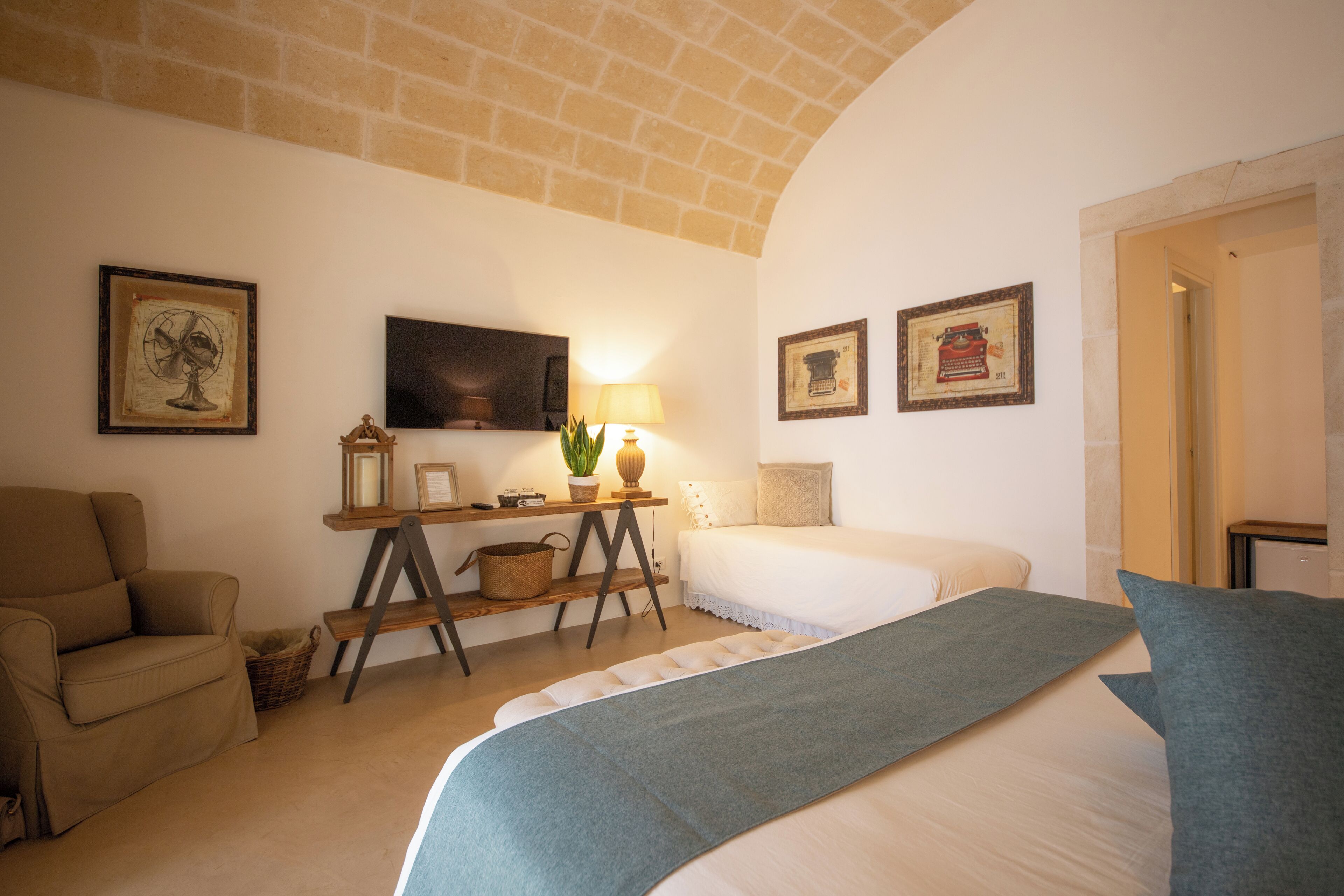 Une pièce chaleureuse avec un plafond en briques, un téléviseur fixé au mur, un canapé, un lit recouvert d’une couverture bleue et des tableaux encadrés accrochés aux murs.