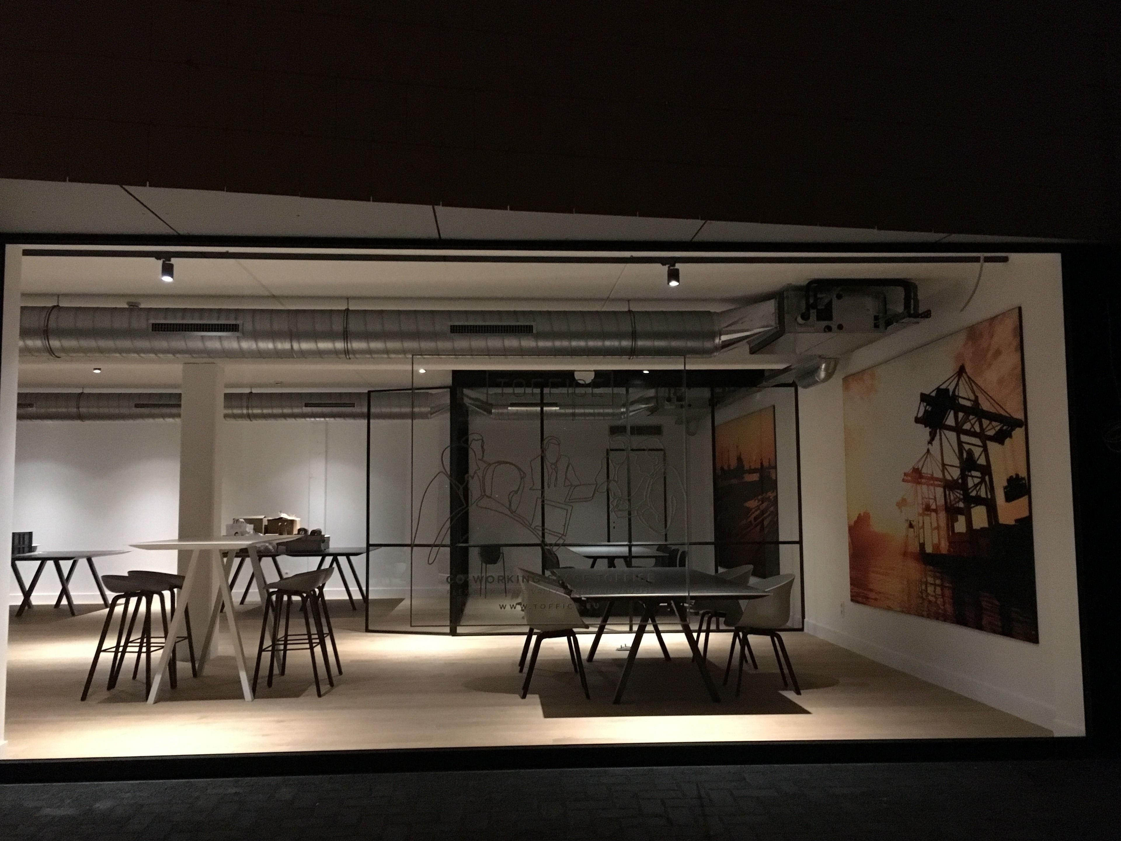 A modern office space with a glass partition, a dining area with contemporary furniture, and a large painting on the wall.