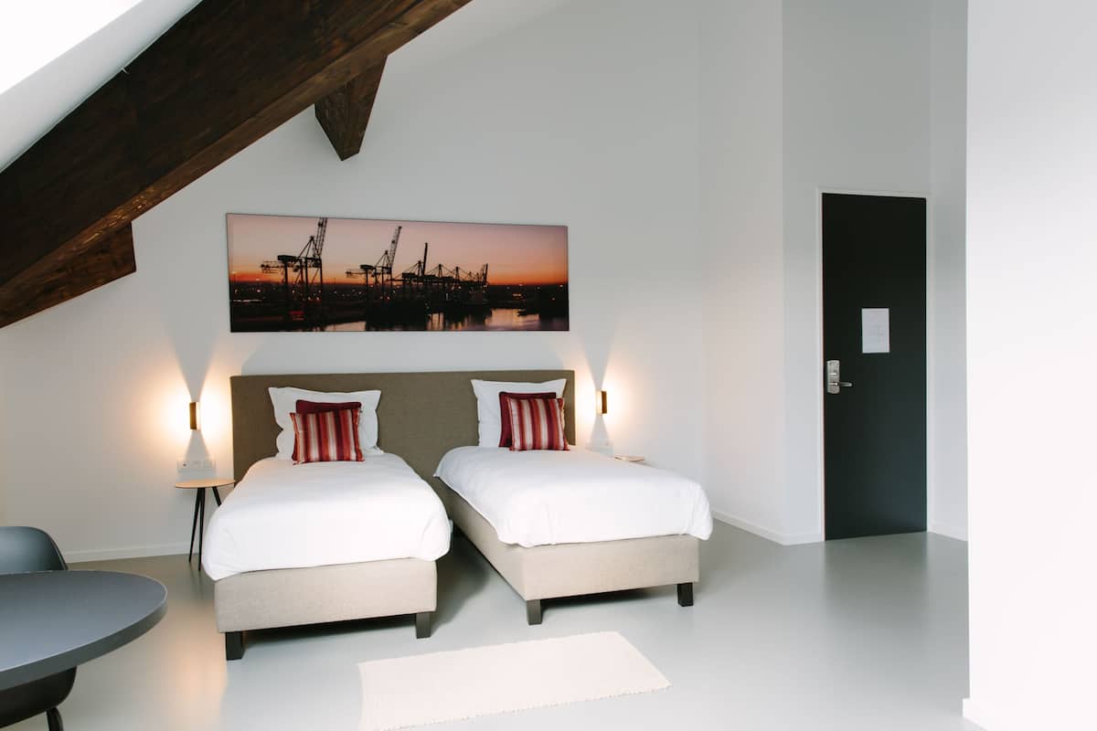 Habitación de hotel con dos camas, un ventanal grande, un cuadro de una puesta de sol y una puerta.