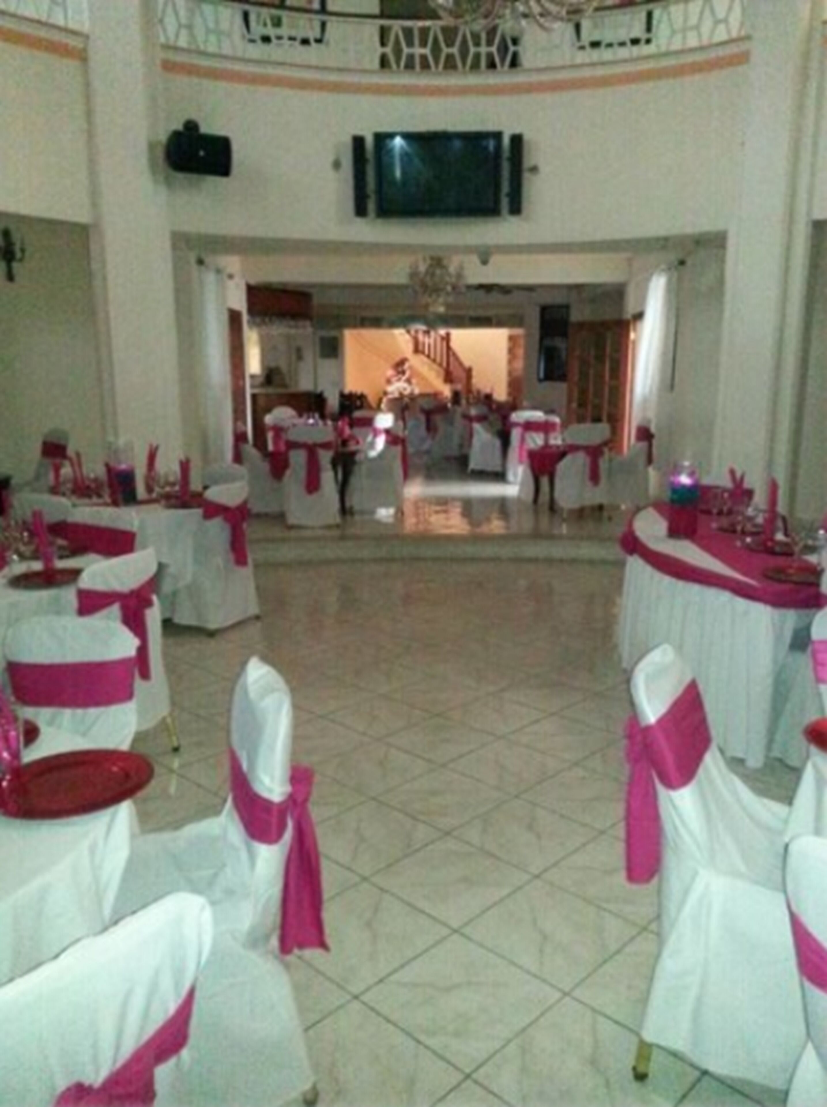 Un salón amplio con mesas y sillas blancas, cintas rosas y una escalera que sube a un nivel superior.