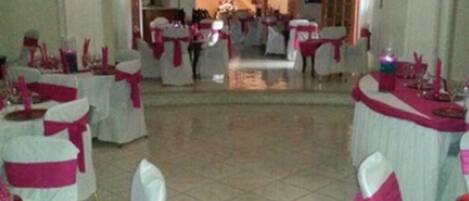 Un salón amplio con mesas y sillas blancas, cintas rosas y una escalera que sube a un nivel superior.