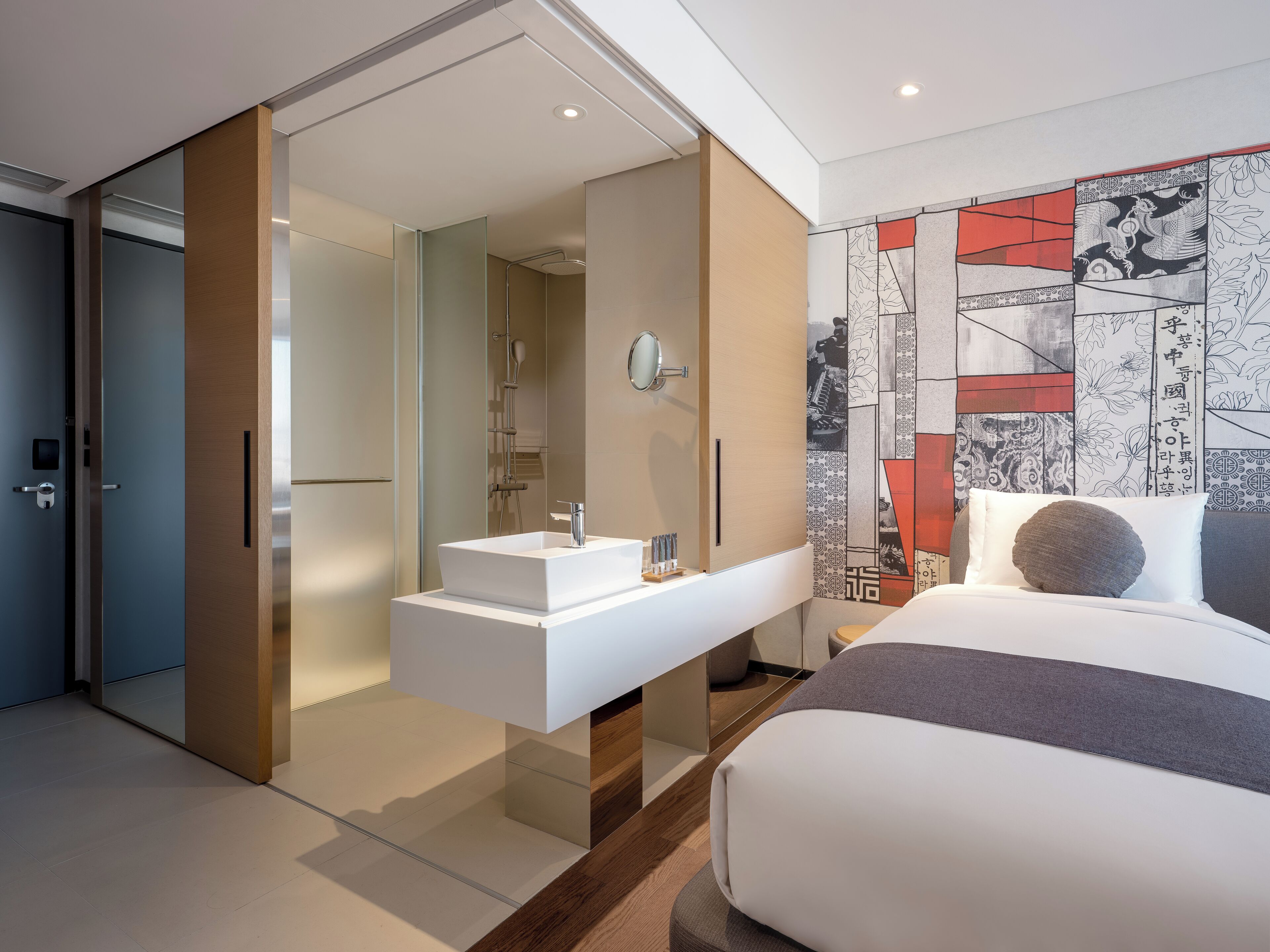 A modern hotel room with a sliding door leading to a bathroom, a built-in white sink, and a large bed with a gray and white color scheme.
