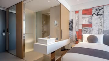 A modern hotel room with a sliding door leading to a bathroom, a built-in white sink, and a large bed with a gray and white color scheme.