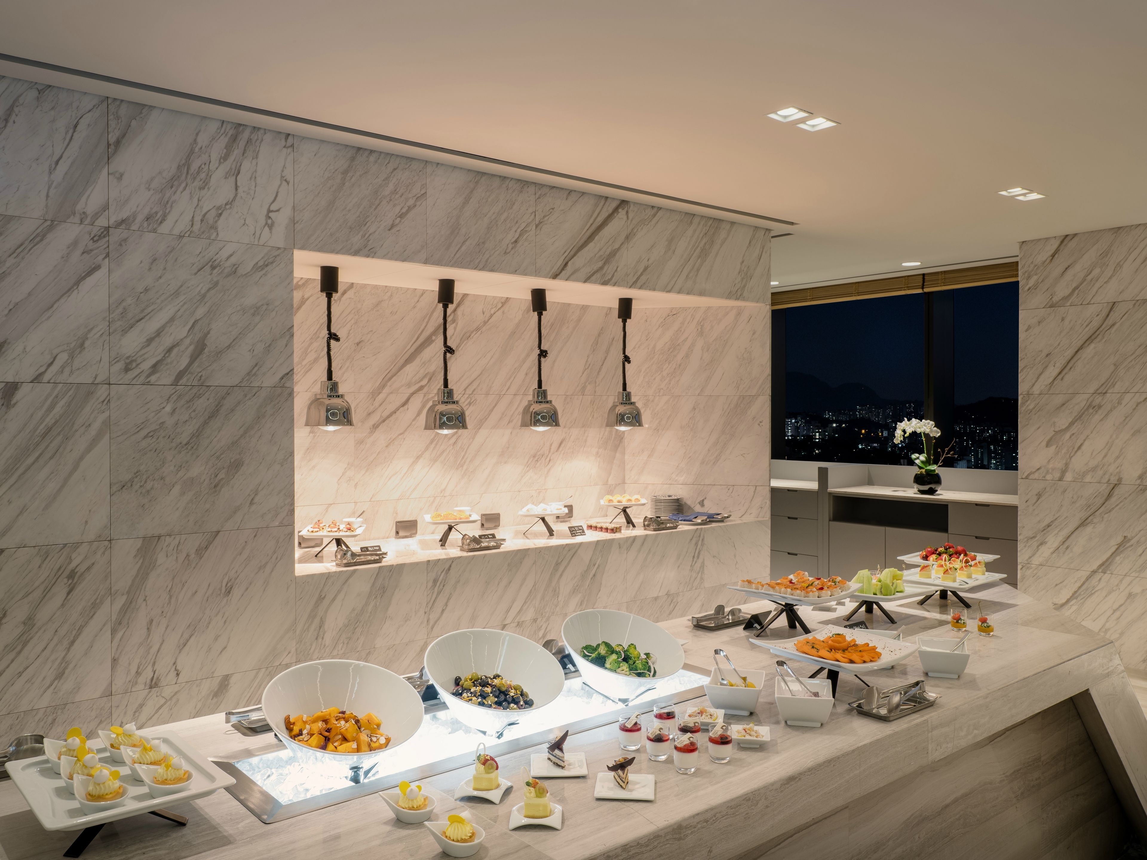 A modern hotel room with a marble wall, a city view, and a buffet with various food items.