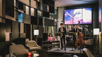 A jazz band performing in a modern hotel lobby with a stage, seating area, and a large screen displaying the band.