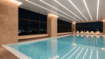 A modern indoor swimming pool with clear blue water, large windows offering city views, and contemporary lighting fixtures.