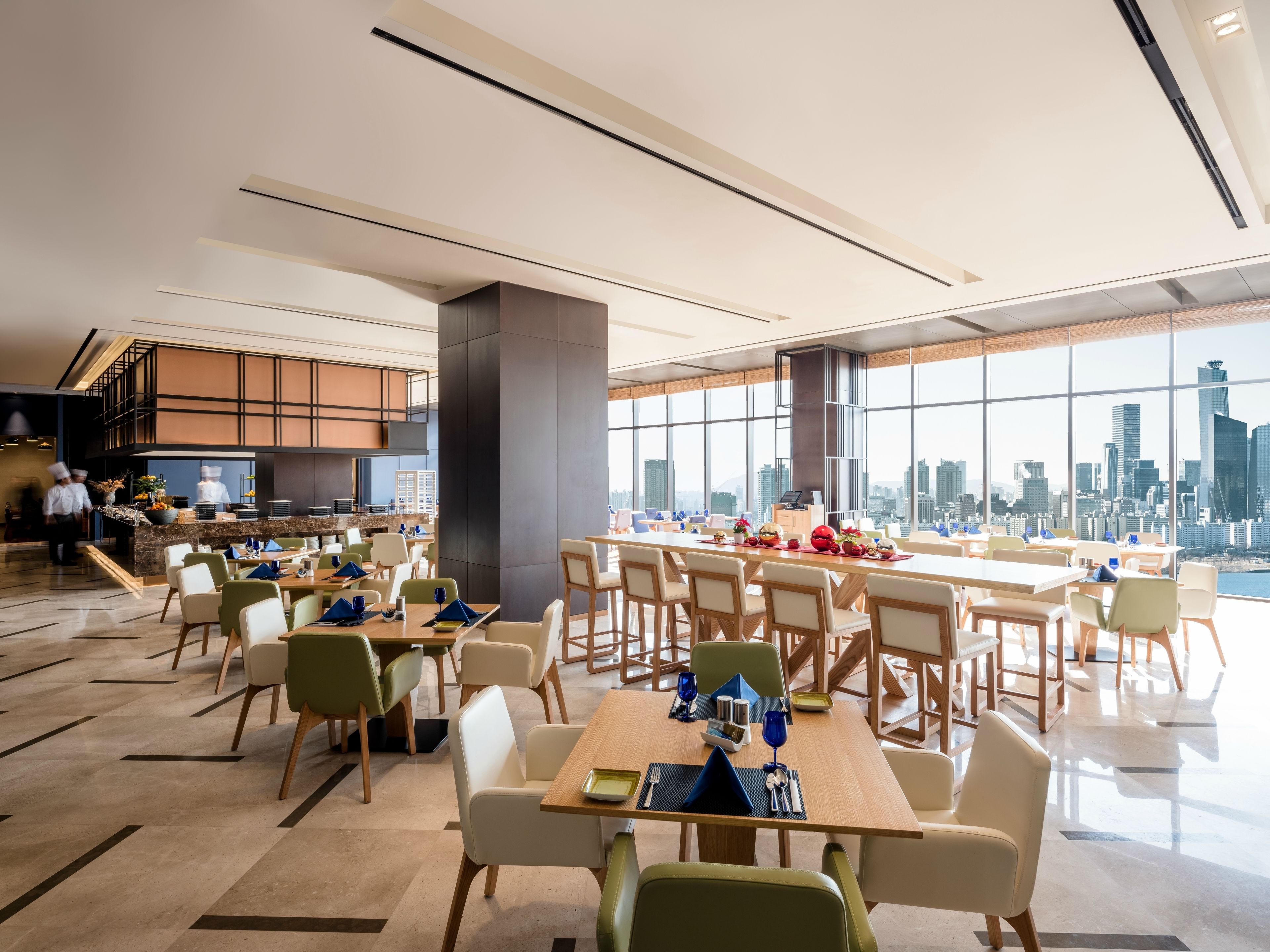 A modern dining area with tables and chairs, a kitchen in the background, and a city view through large windows.