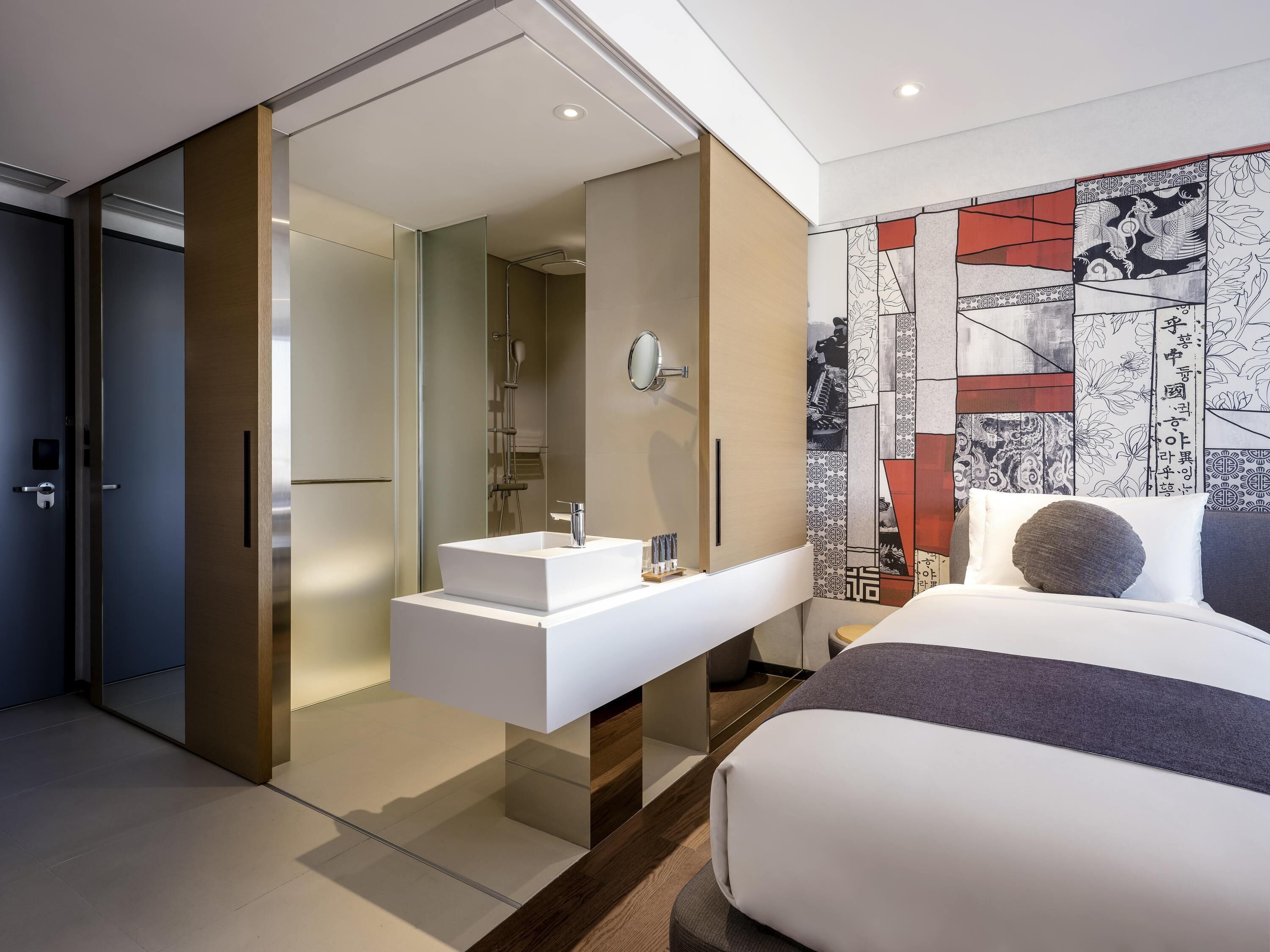 A modern hotel room with a glass-enclosed shower, a white vanity with a sink, and a large bed with a gray and white color scheme.