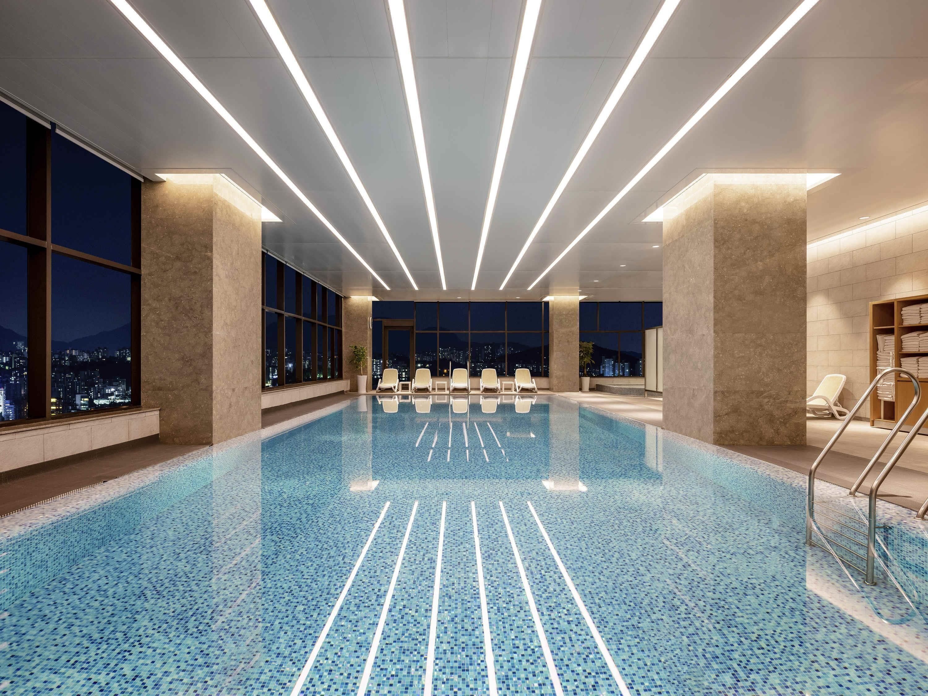 A modern indoor swimming pool with a city view, large windows, and a clean, tiled floor.