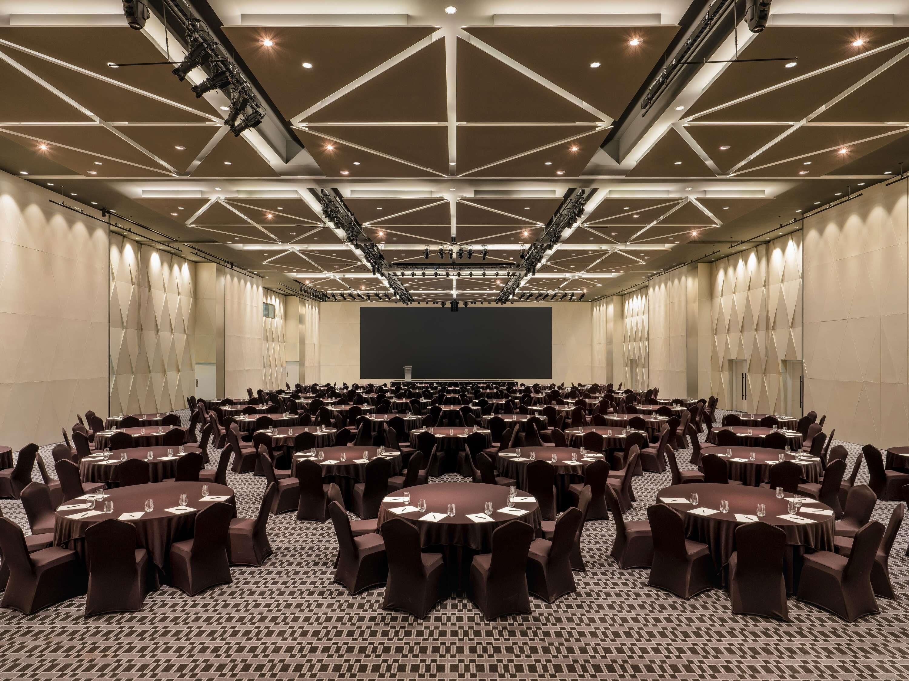 A large conference room with multiple round tables and chairs, a stage with a large screen, and a high ceiling with geometric lighting.
