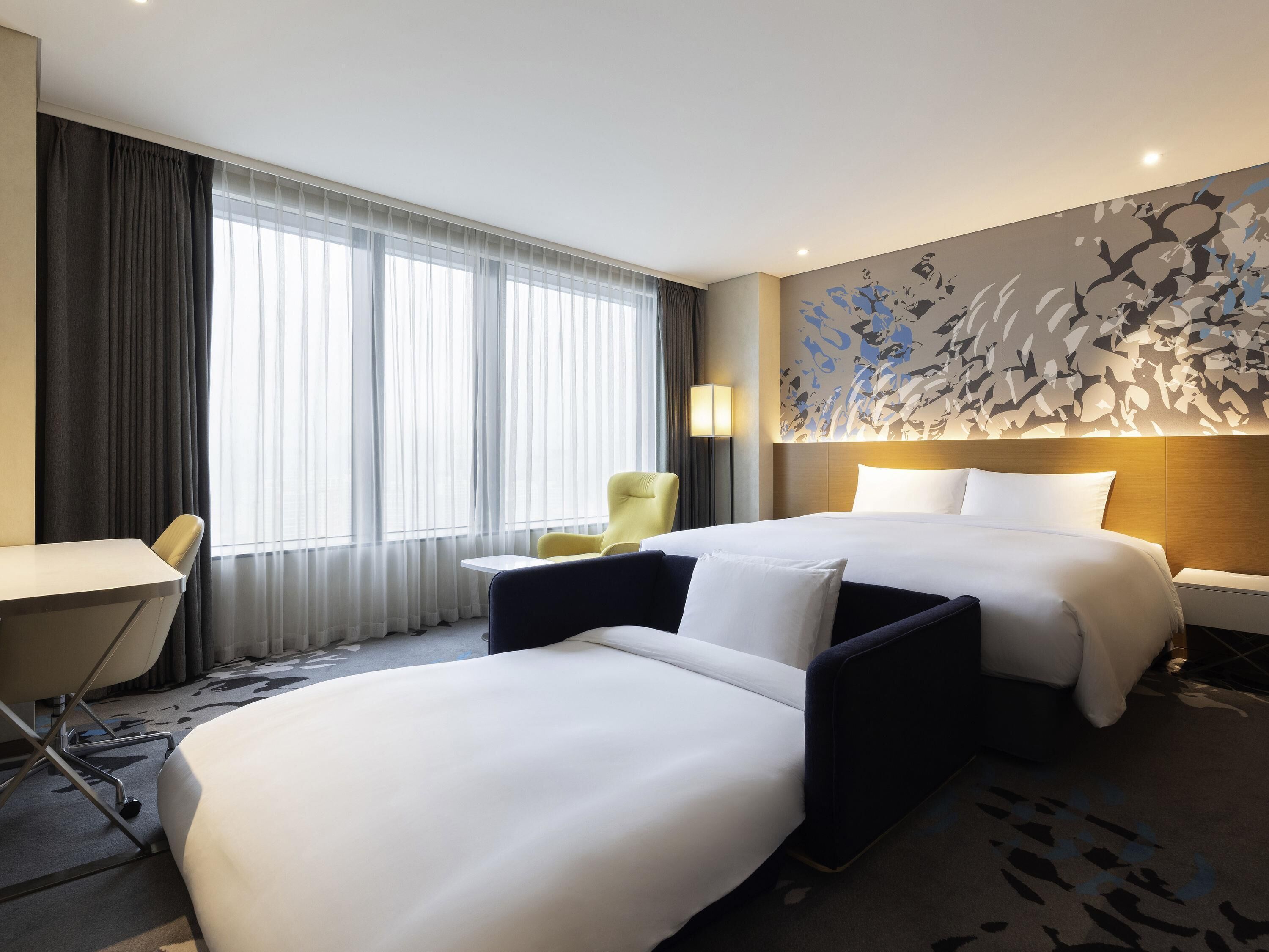 A hotel room with two beds, a desk, a chair, and a large window with curtains.