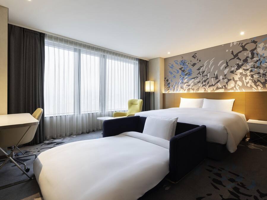 A hotel room with two beds, a desk, a chair, and a large window with curtains.