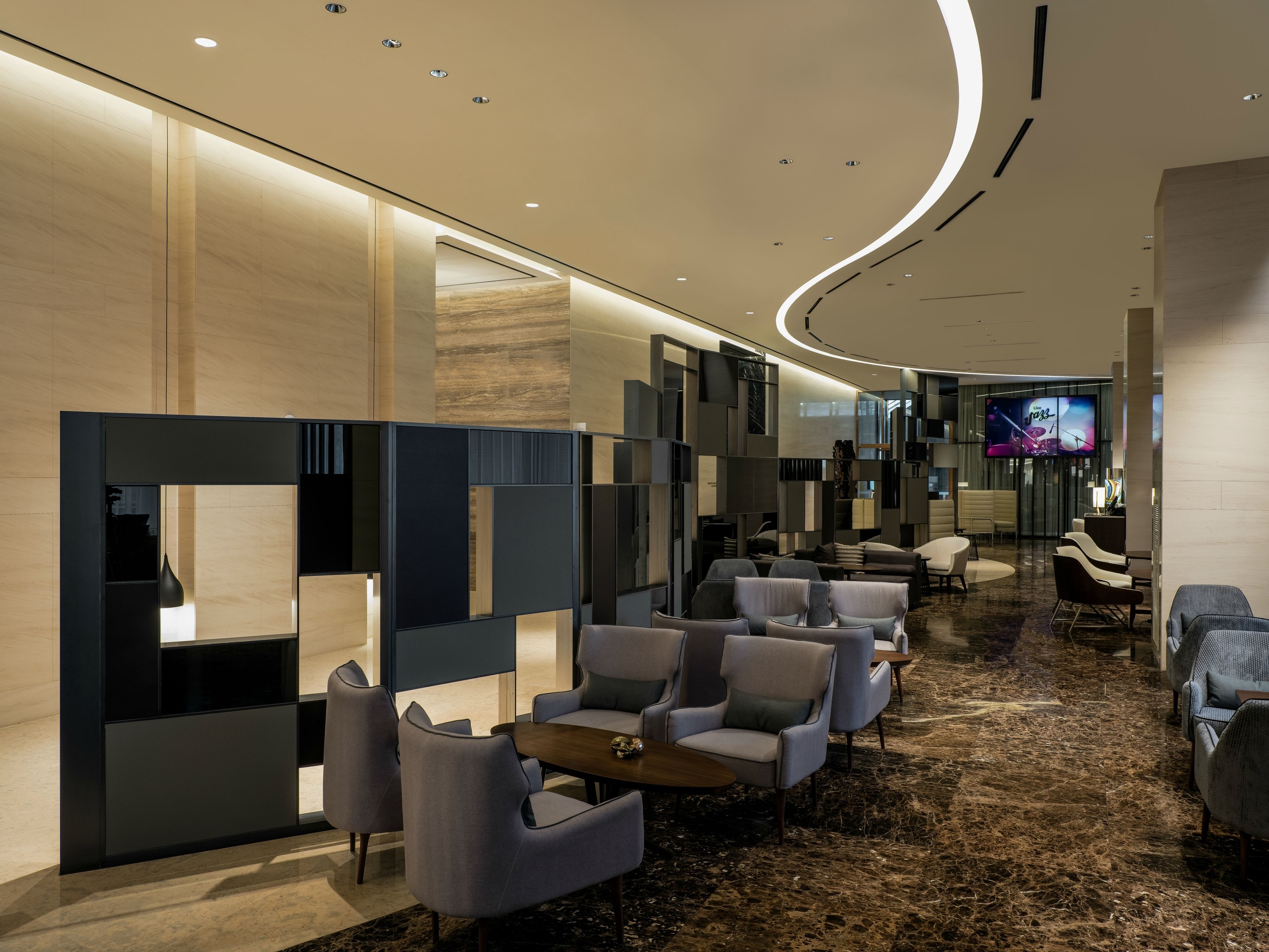 A modern hotel lobby with a curved ceiling, seating area with sofas and tables, and a television screen displaying a jazz program.