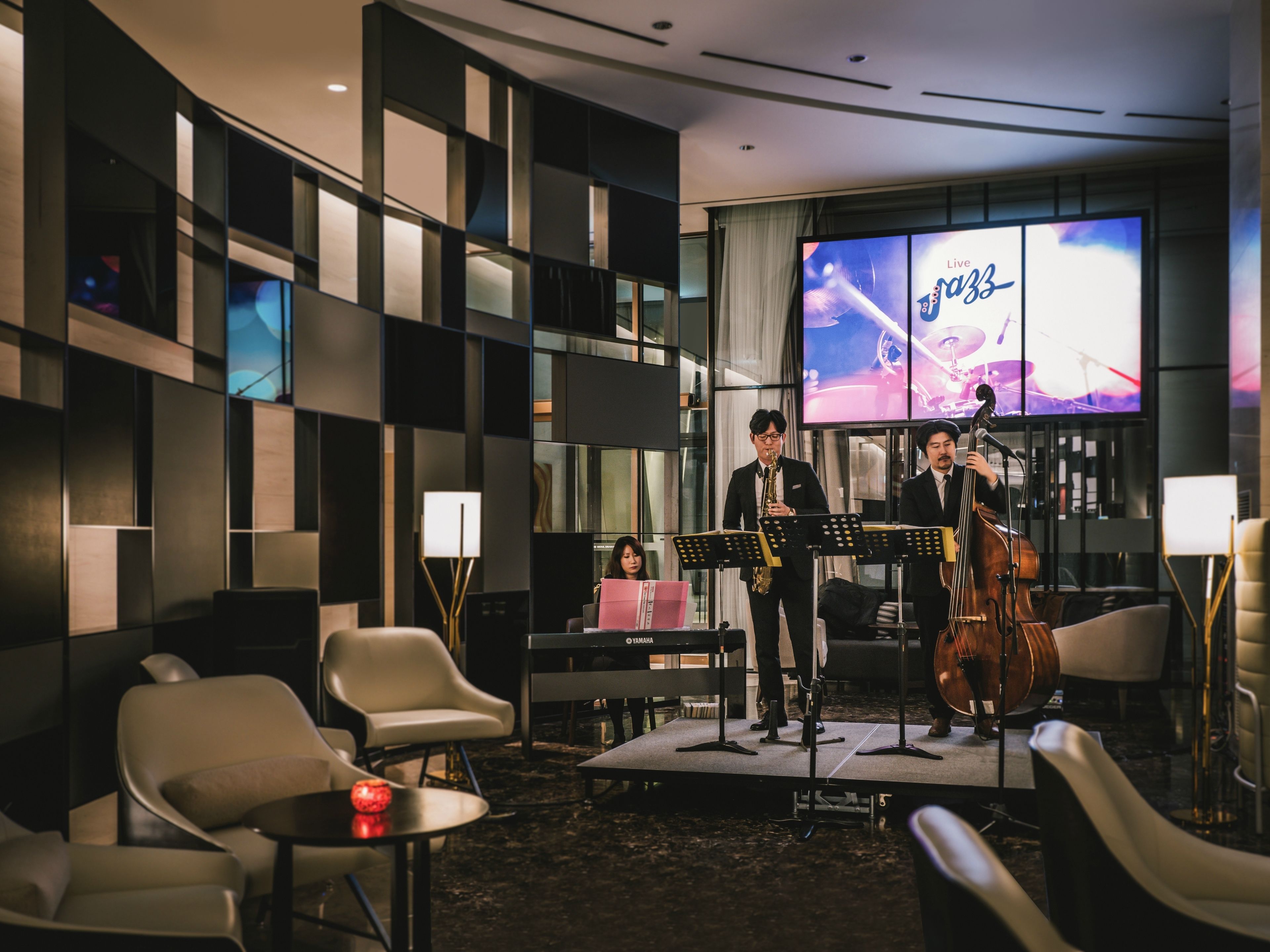 A jazz band performing in a modern hotel lobby with a stage, seating area, and a large screen displaying the band.