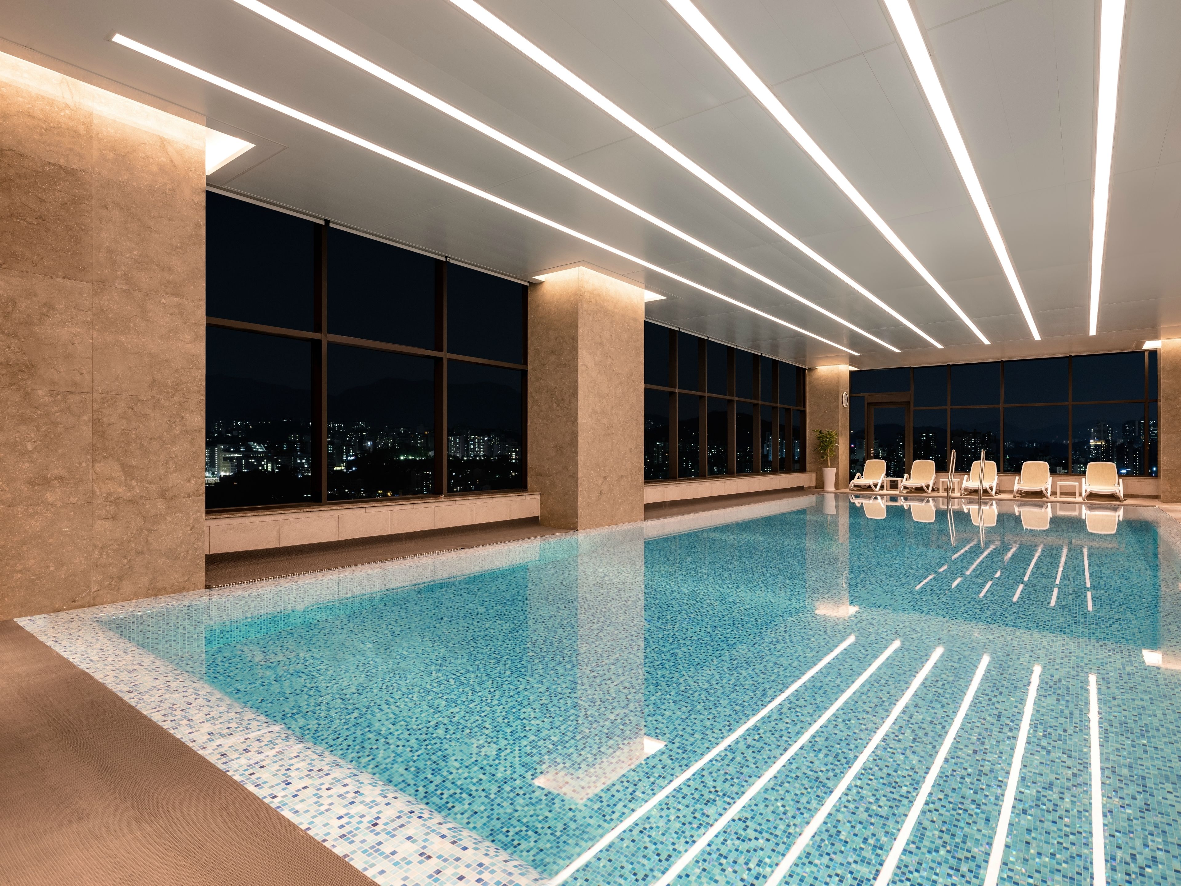 A modern indoor swimming pool with clear blue water, large windows offering city views, and contemporary lighting fixtures.