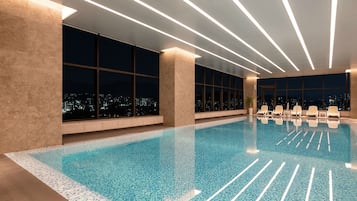 A modern indoor swimming pool with clear blue water, large windows offering city views, and contemporary lighting fixtures.