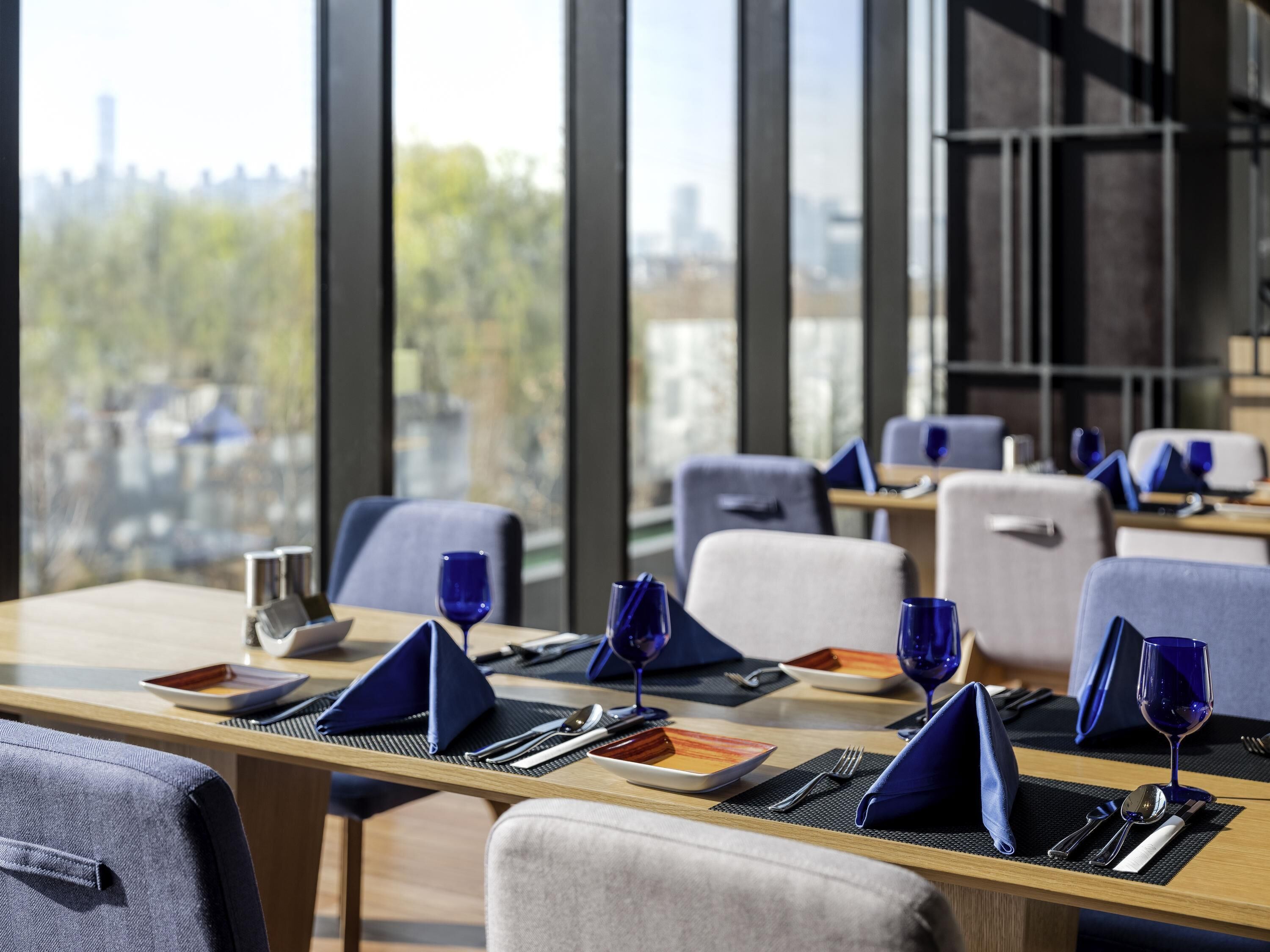 A modern dining area with tables set for two, featuring blue napkins, wine glasses, and cutlery. Large windows offer a view of the cityscape.